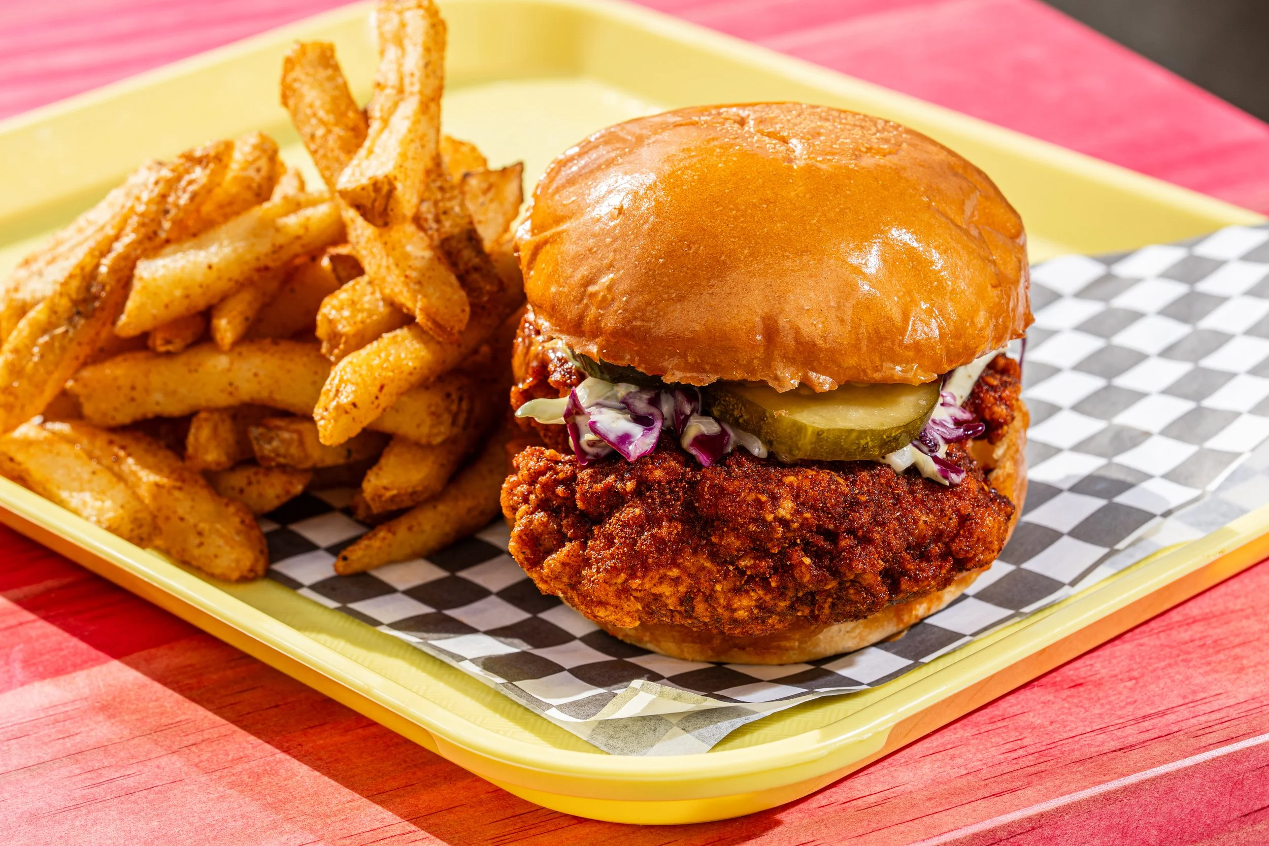 Fried chicken sandwich with pickles and coleslaw on a bun next to seasoned French fries on a yellow tray.
