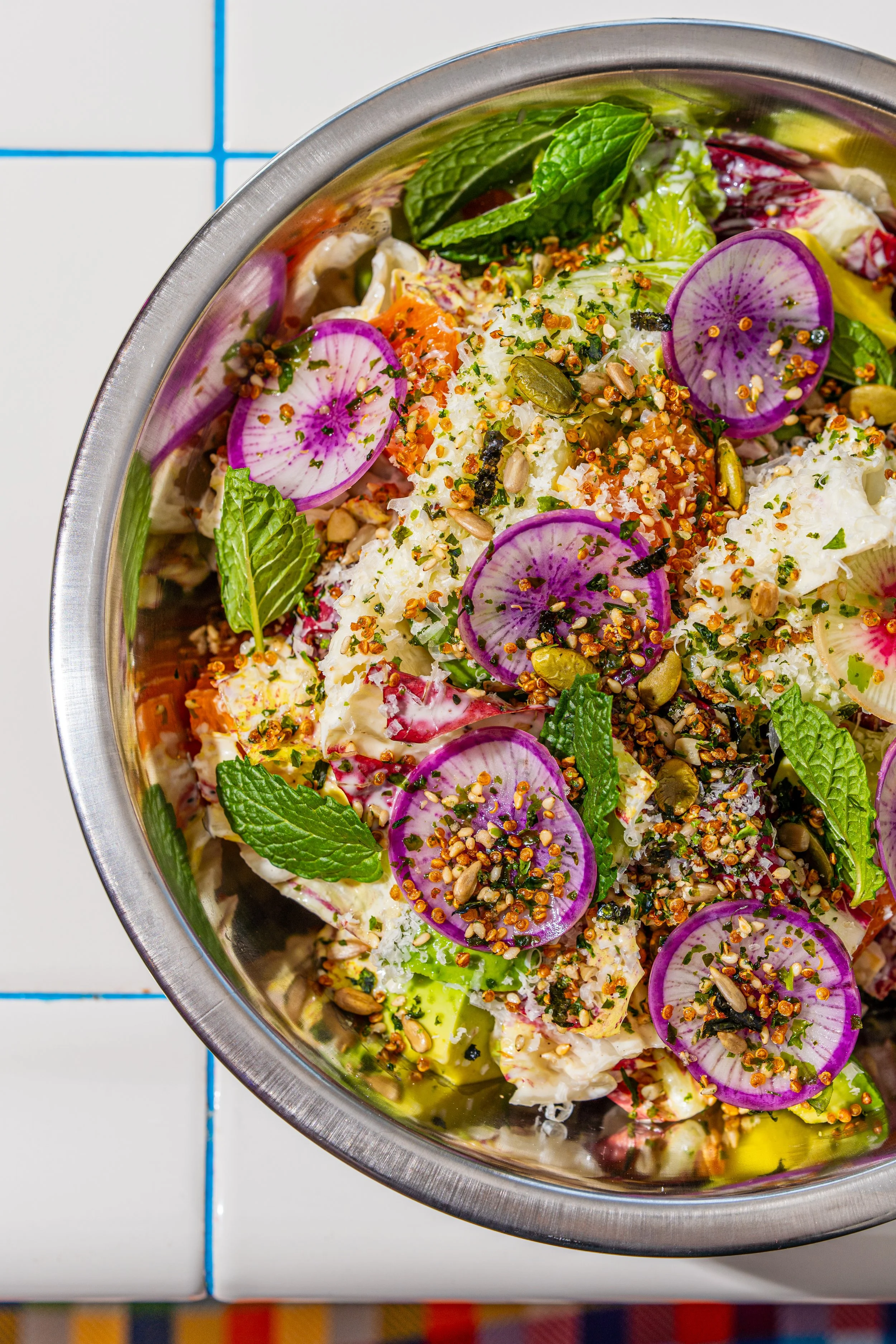 A close-up of a fresh salad in a metal bowl, containing sliced radishes, mint leaves, chopped herbs, seeds, and vegetables.