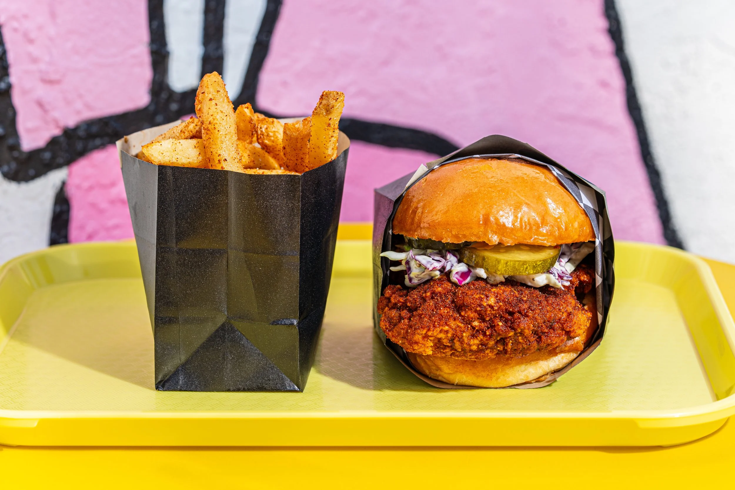 Fried chicken sandwich with pickles and coleslaw in a brioche bun, served with seasoned French fries on a yellow tray in front of a pink and black graffiti background.