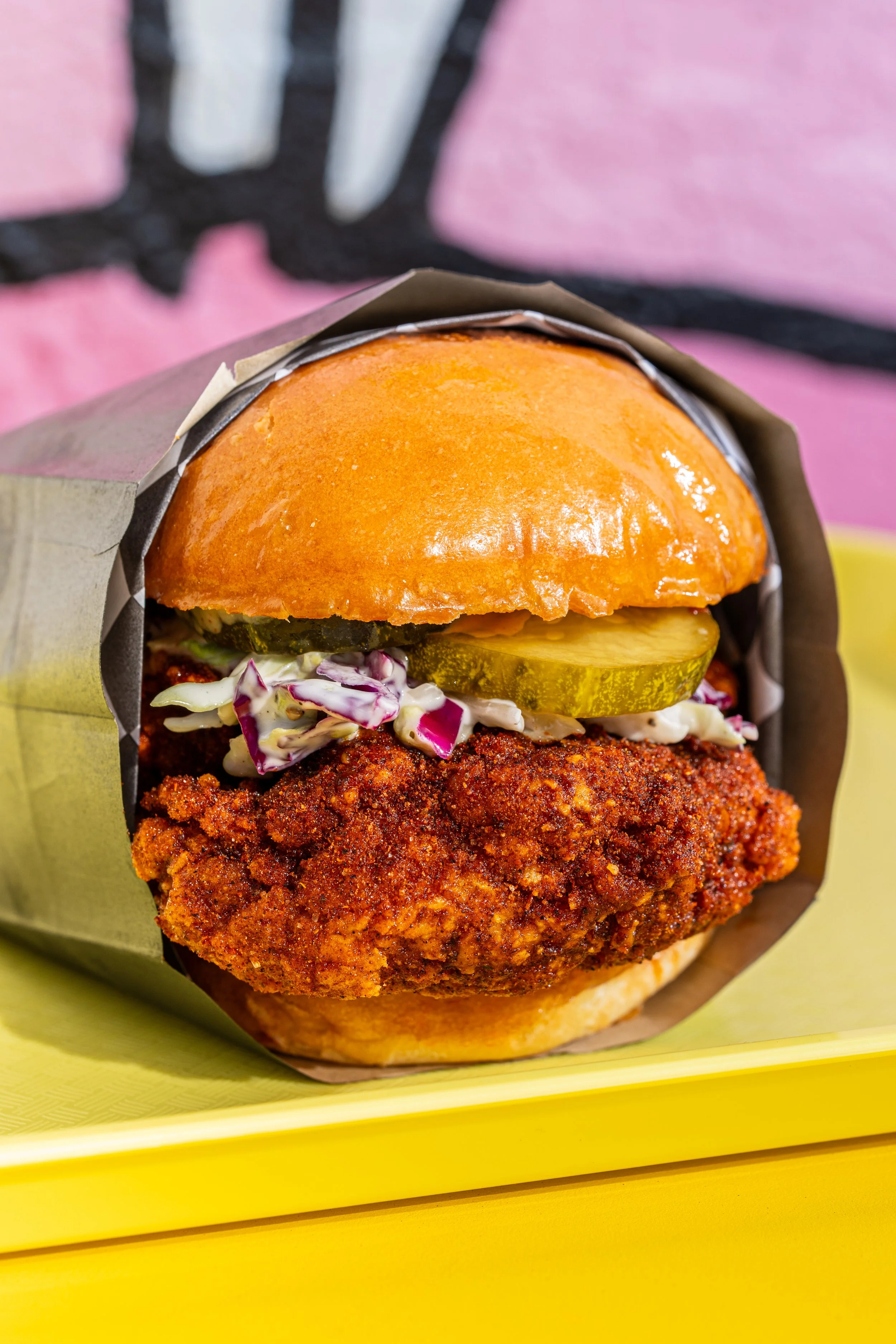 Close-up of a fried chicken sandwich with pickles, coleslaw, and a brioche bun, on a yellow tray.