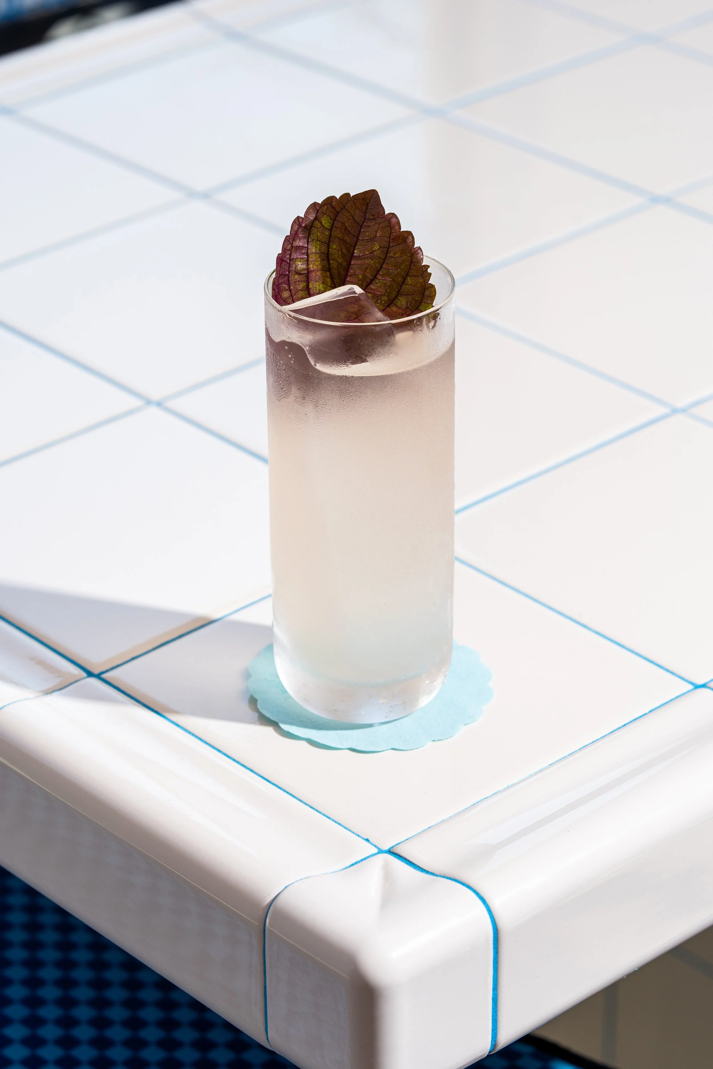 A tall glass of a layered cocktail garnished with purple-leafed herbs resting on a white tiled surface