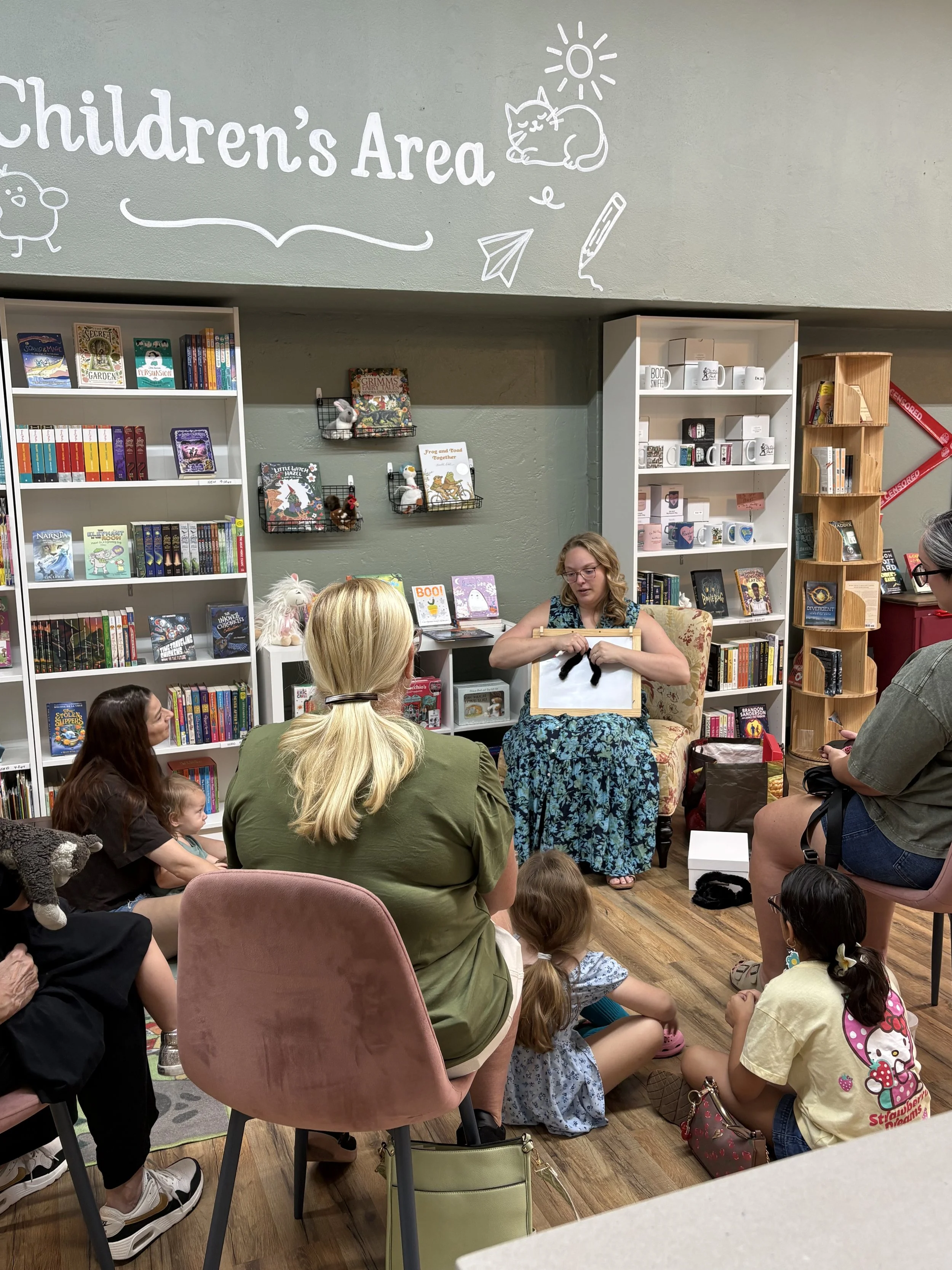 A woman is teaching a puppet show to children sitting on the floor with their attention focused on her, in a bookstore or library setting with shelves of books and stuffed animals around.