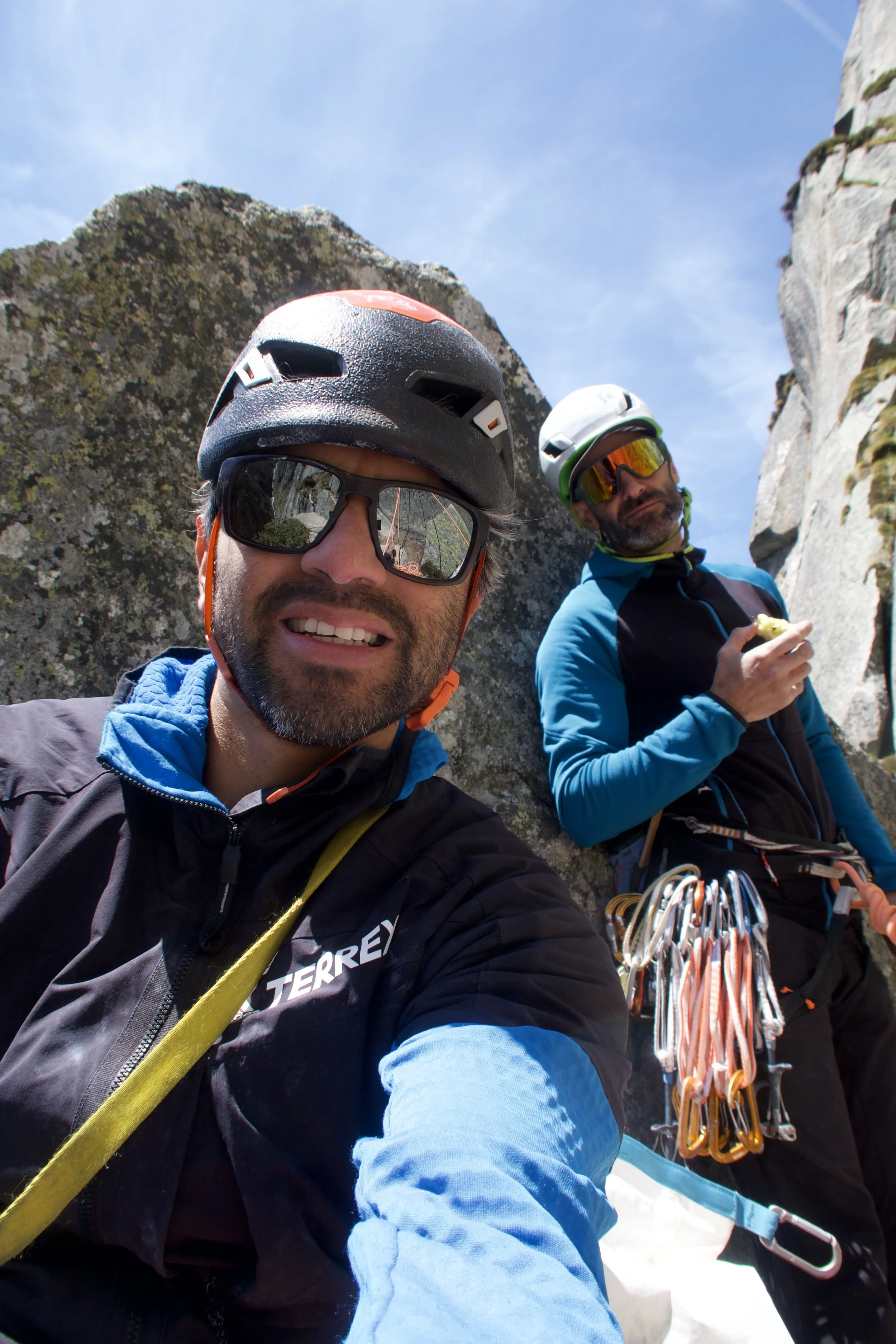 Zwei Bergsteiger in Kletterausrüstung und Helmen vor einer Felswand, einer macht ein Selfie, während der andere eine Banane isst, bei Sonnenlicht unter blauem Himmel.