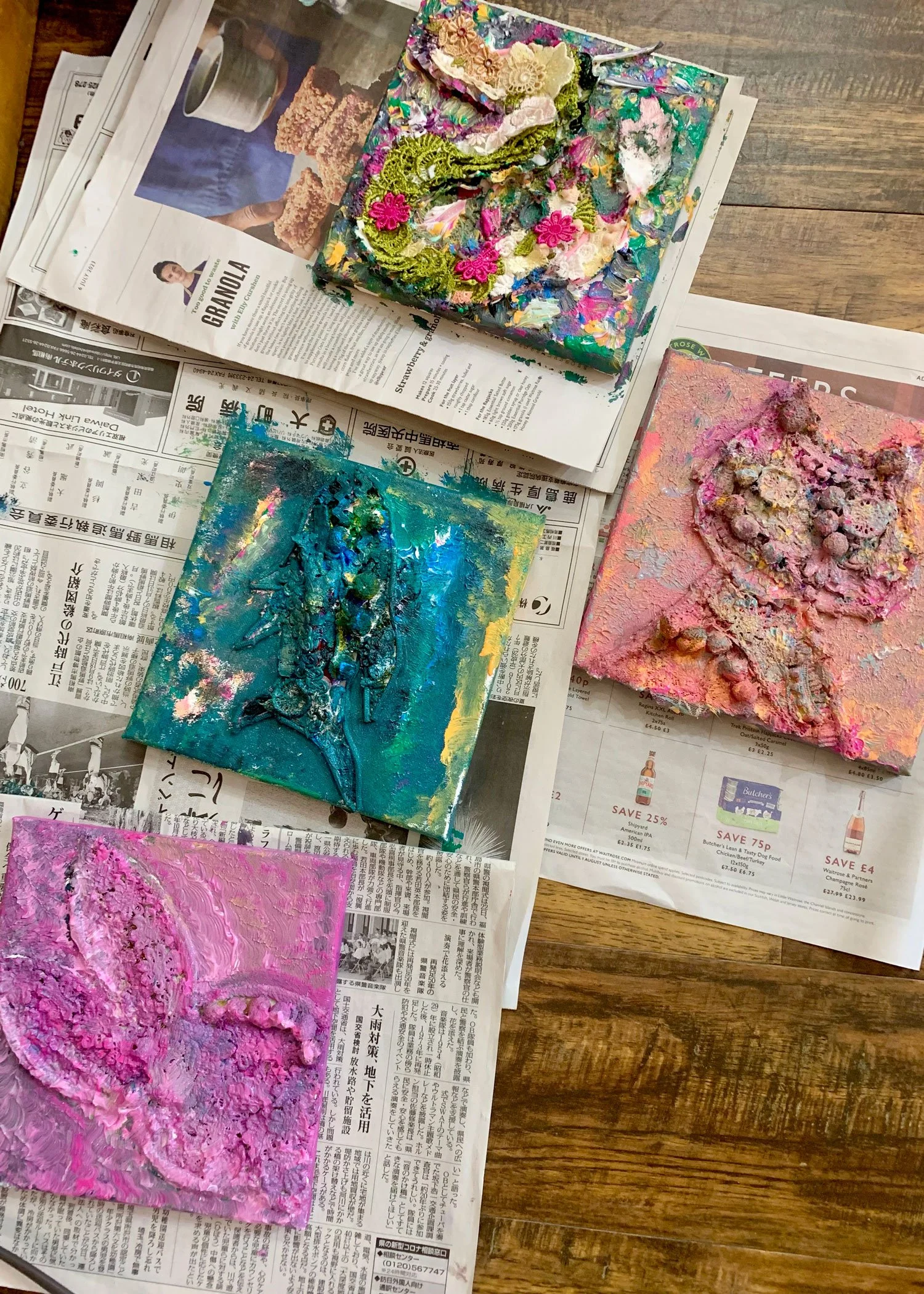 Painted canvases on newspaper-covered wooden floor, featuring abstract and textured artwork in various colors including pink, blue, green, and cream.