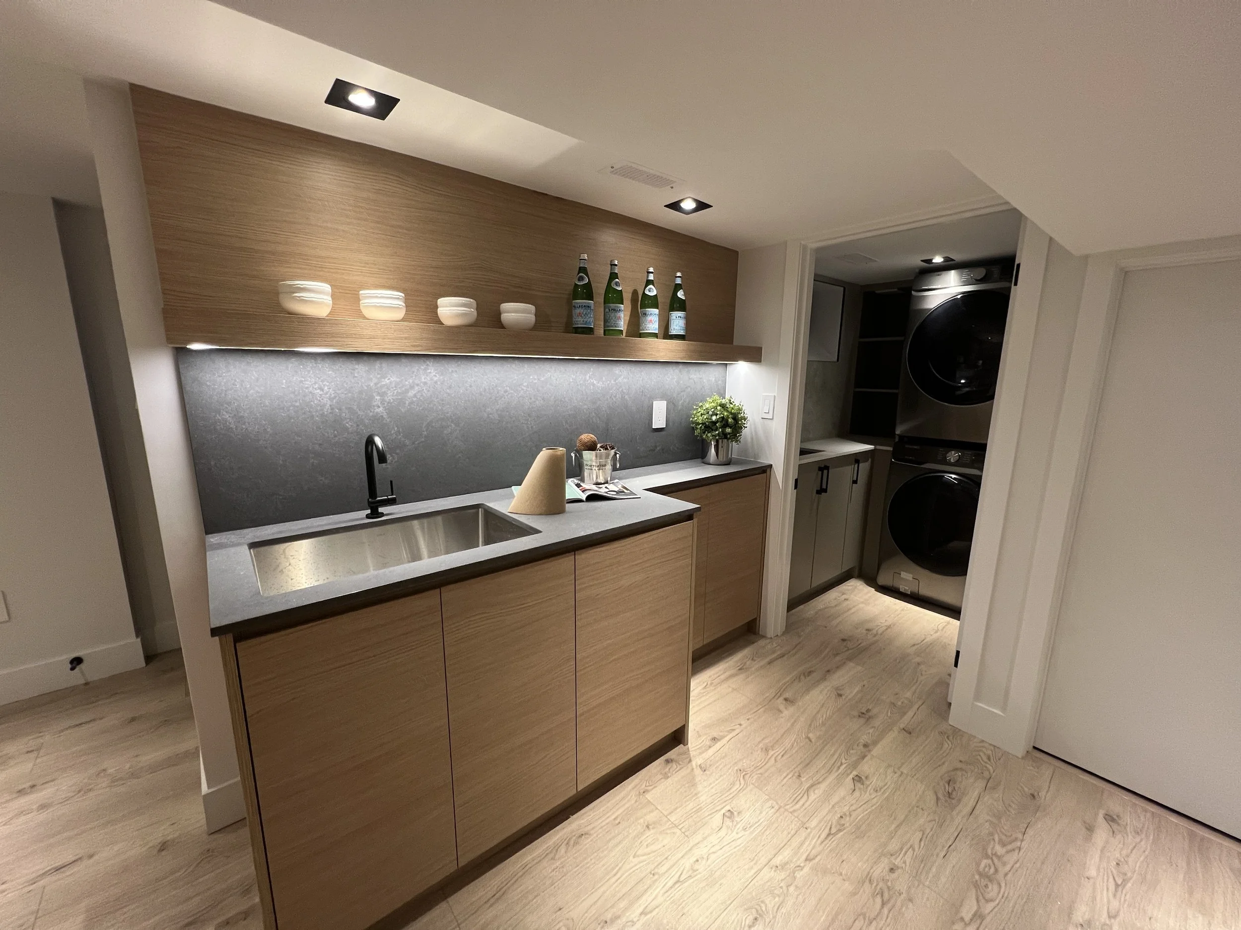 Modern laundry area with wooden cabinetry, black countertop, sink, potted plant, and stacked washer and dryer.