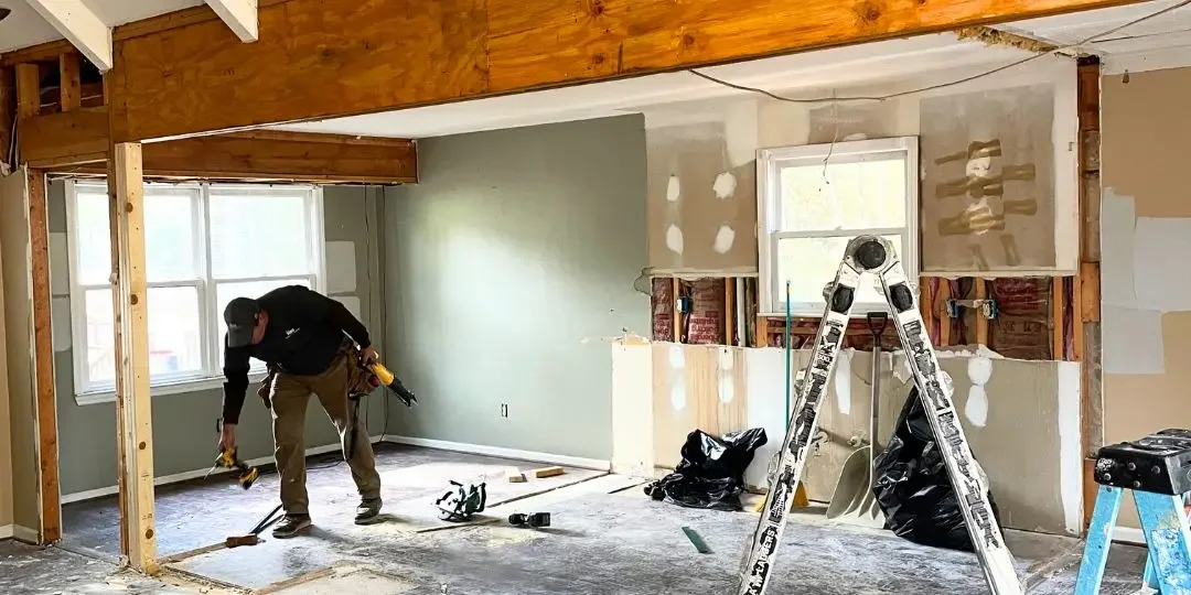 A person is working on a home renovation project inside a room, using tools to work on the floor and walls. The room has windows, partially finished walls, and construction materials.