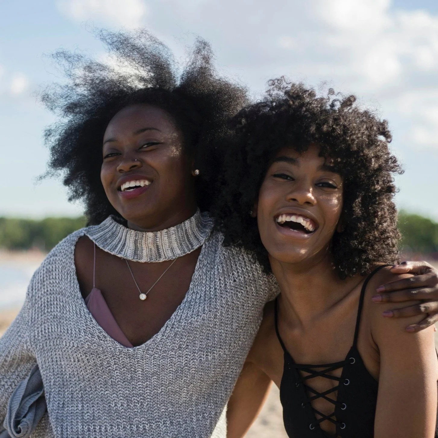Two women with curly hair smiling and embracing outdoors on a beach or lakeside, with water and trees in the background.