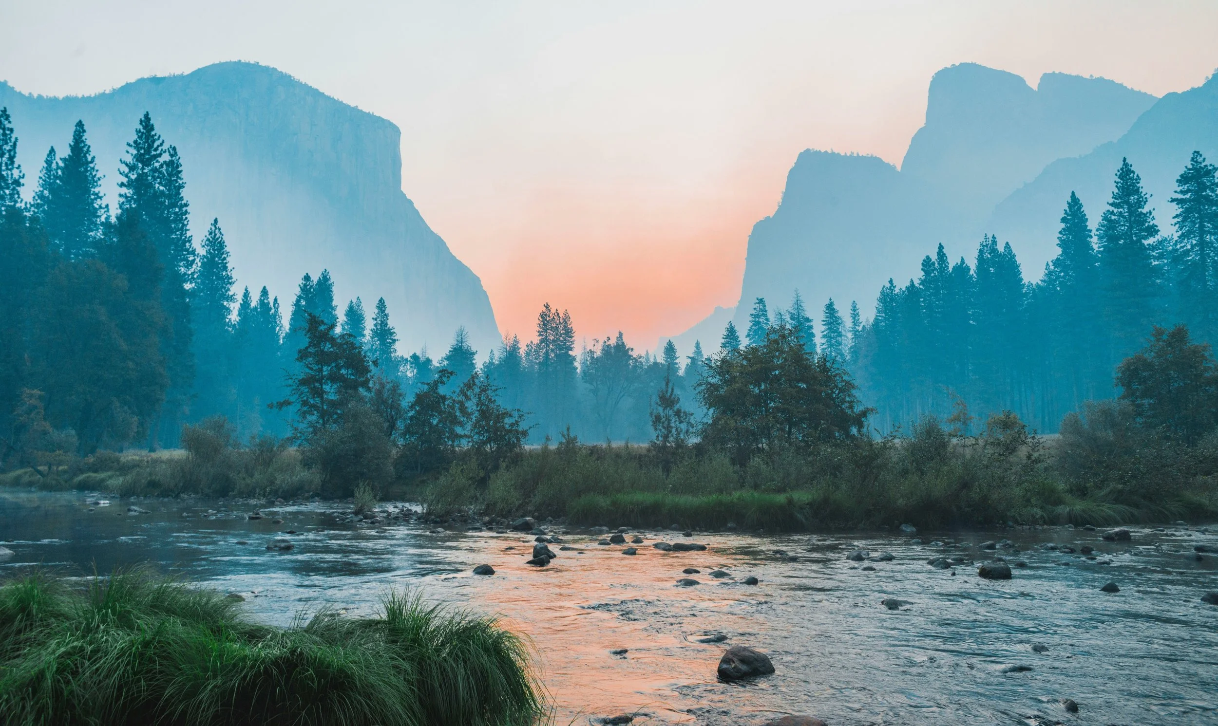 A scenic view of a river flowing through a forest with tall pine trees, mountains in the background, and a pastel-colored sky at either sunrise or sunset.