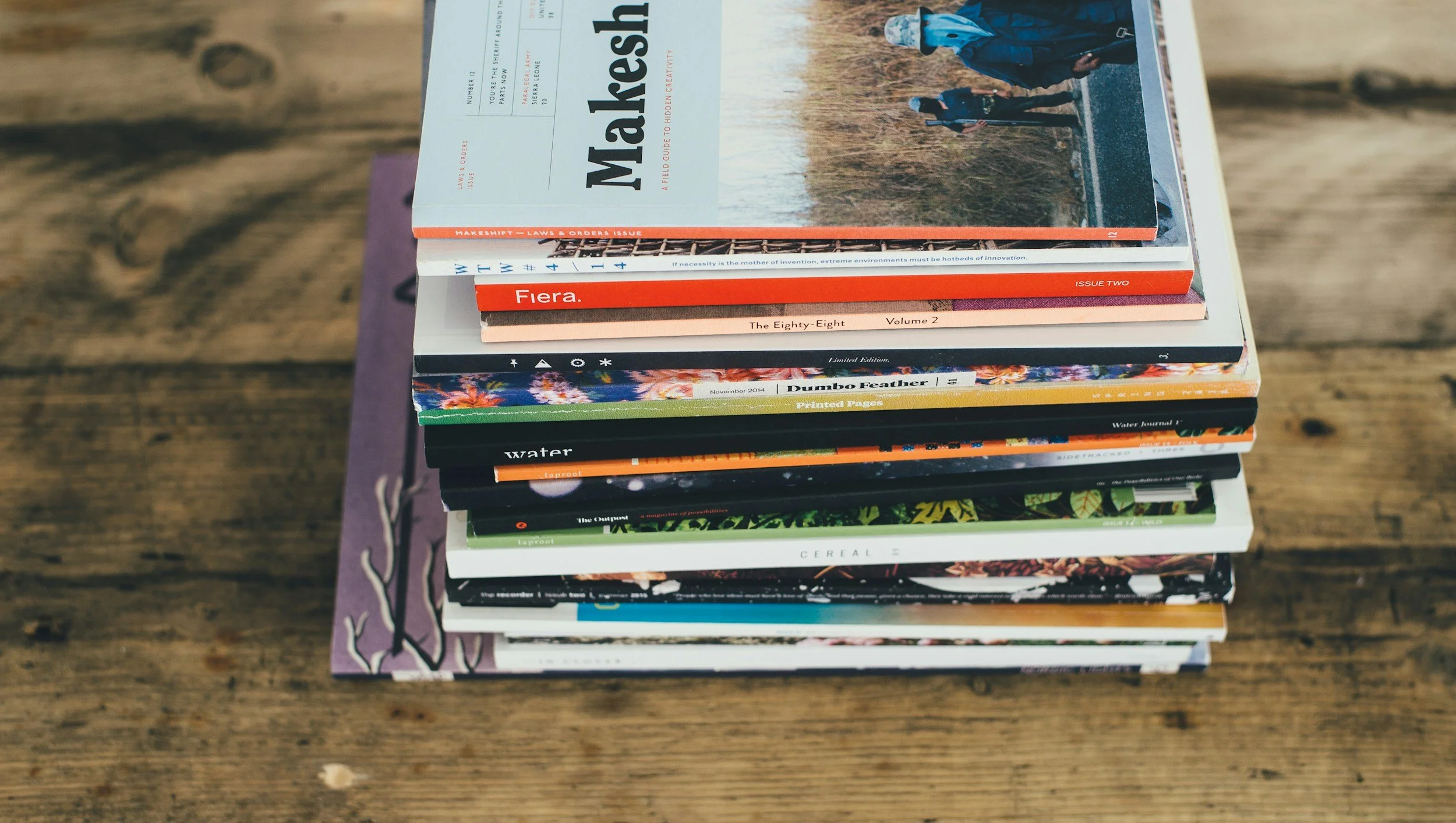 Stack of various magazines and books placed on a wooden surface, including titles like 'Makershift', 'Fiera', 'The Eighty-Eight Volume 2', and others with colorful covers.