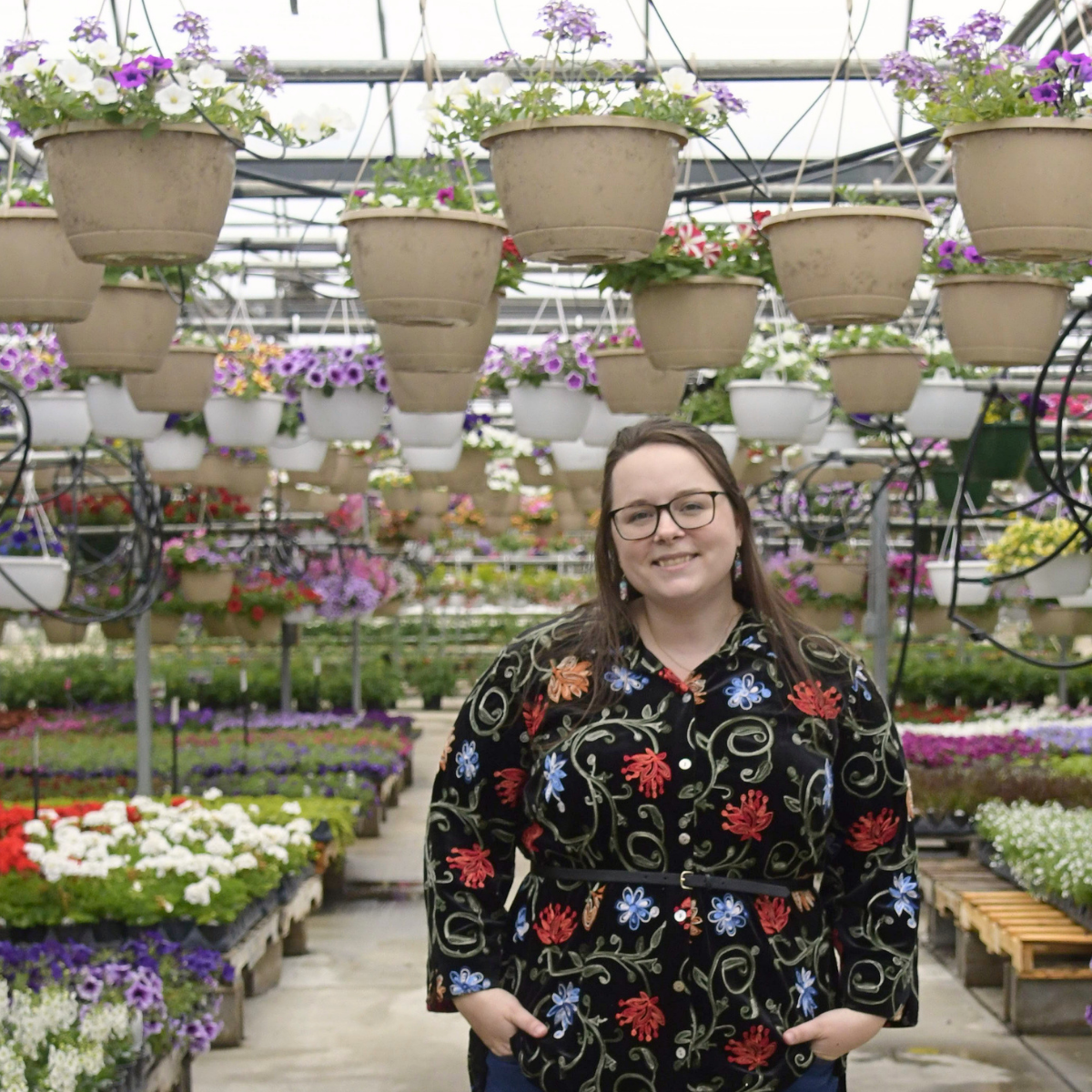 Smiling woman with brown hair and glasses stands in vibrant greenhouse surrounded by flowers