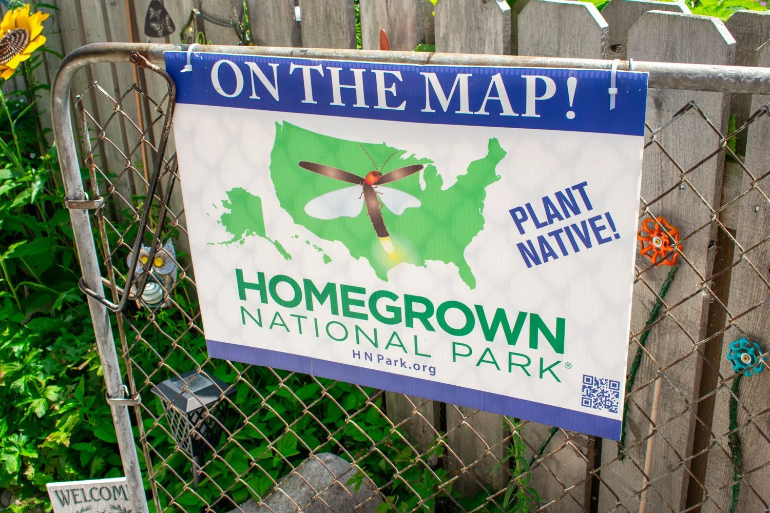 A large sign stating "Homegrown National Park" is attached to a chainlink fence, surrounded by flowers and outdoor decor