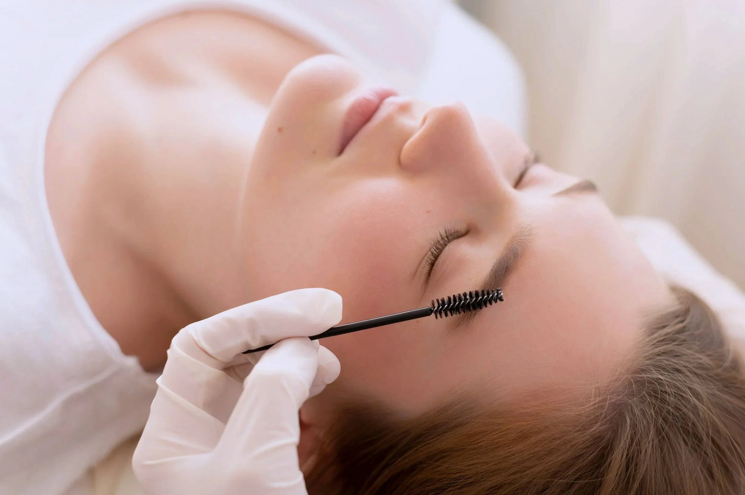 A woman with closed eyes lying on a bed during a beauty treatment, while a person with a gloved hand uses a spoolie brush on her eyelashes.