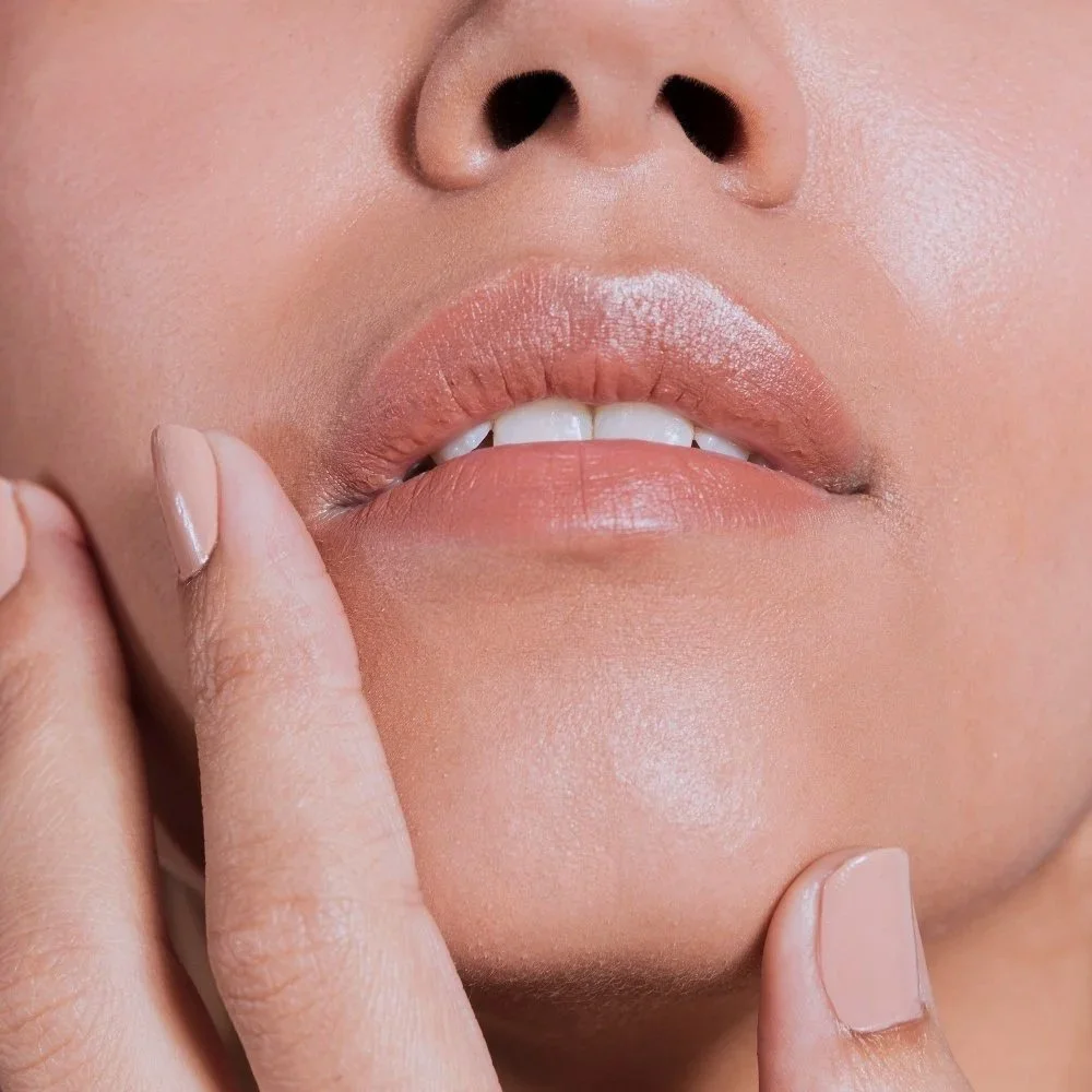 Close-up of a woman's lips with fingers touching her chin, showing natural makeup, slightly parted lips, and a finger touching her cheek.