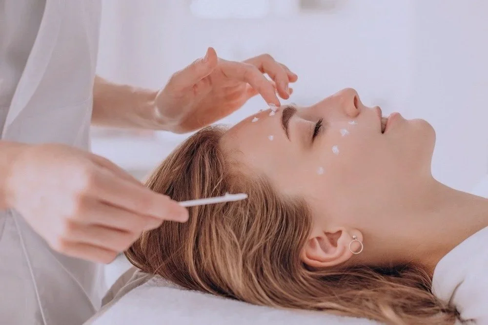 A woman lying down during a facial treatment, with a skincare professional applying cream or serum to her forehead.