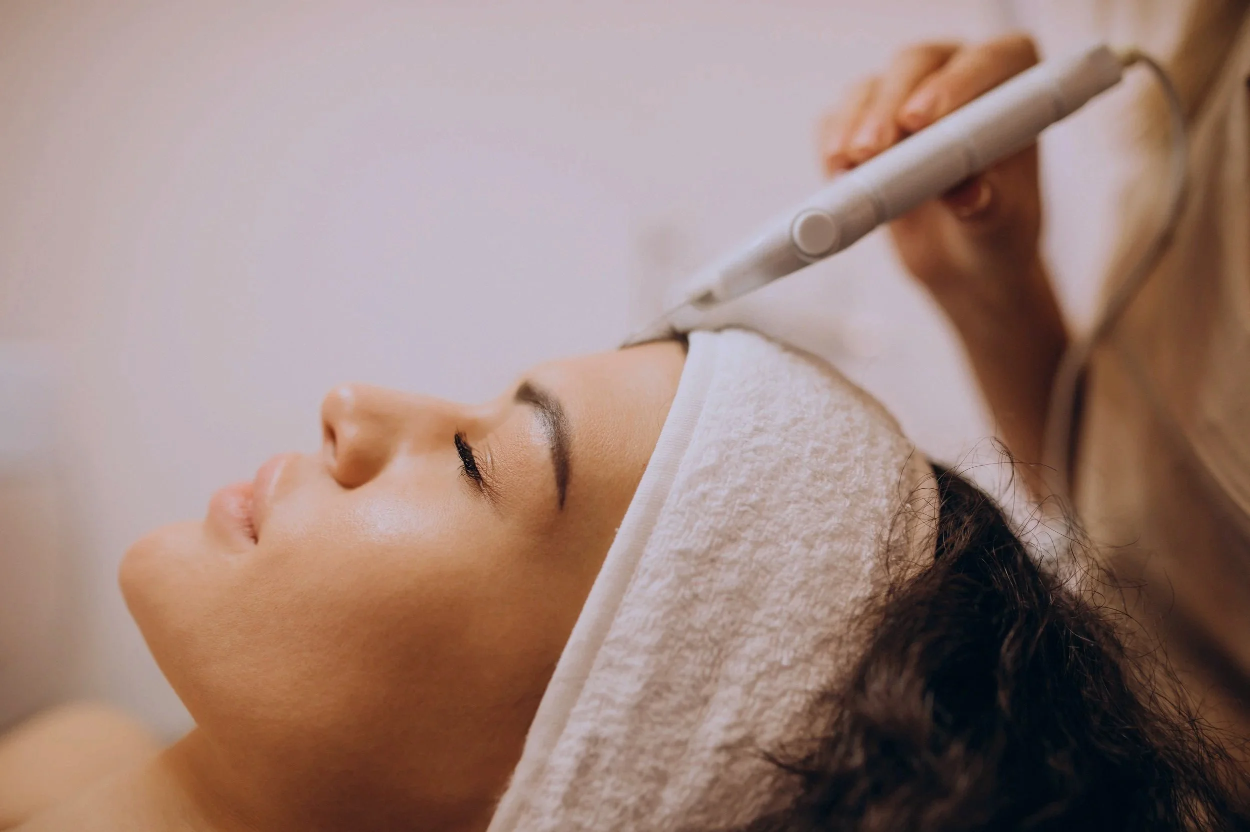A woman receiving a facial treatment, lying down with her eyes closed, while a professional applies a treatment with a handheld device.