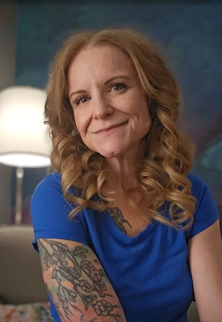 A woman with curly red hair, wearing a blue shirt, smiles at the camera. She has tattoos on her arm and chest, and is seated indoors with a lamp and abstract art in the background.