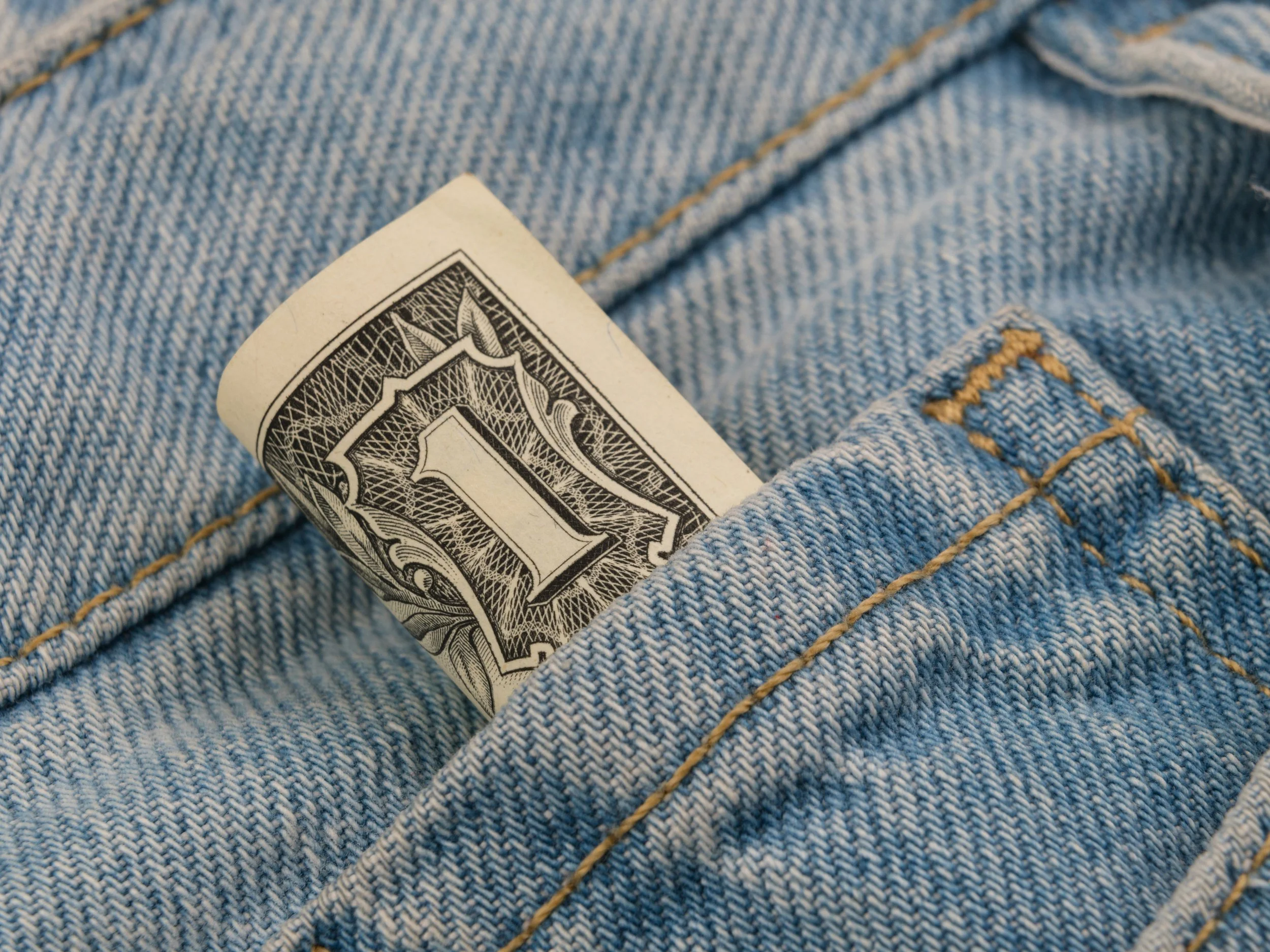 Close-up of a folded pair of blue denim jeans with a one-dollar bill partially tucked into the front pocket.
