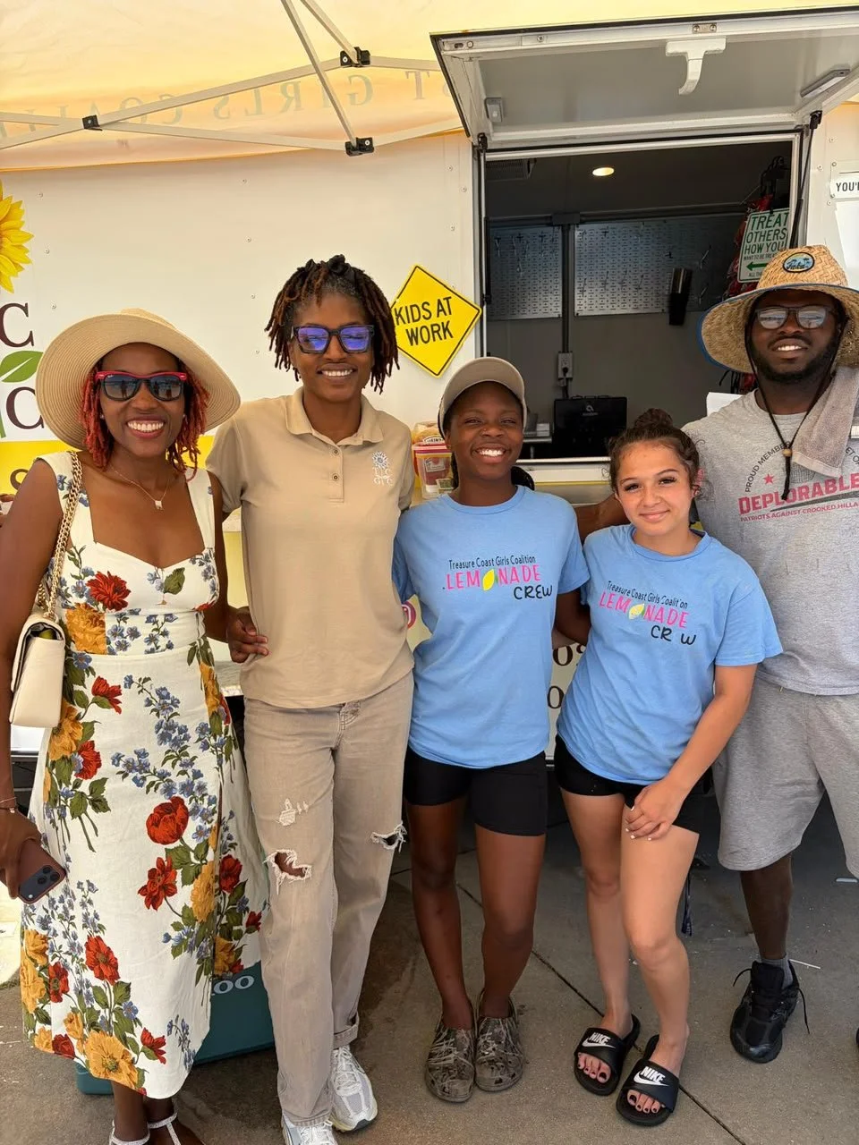 Group of five people standing in front of a food truck at an outdoor event, smiling and posing for the camera. Two women are wearing hats and sunglasses, and two young girls are wearing matching blue shirts.