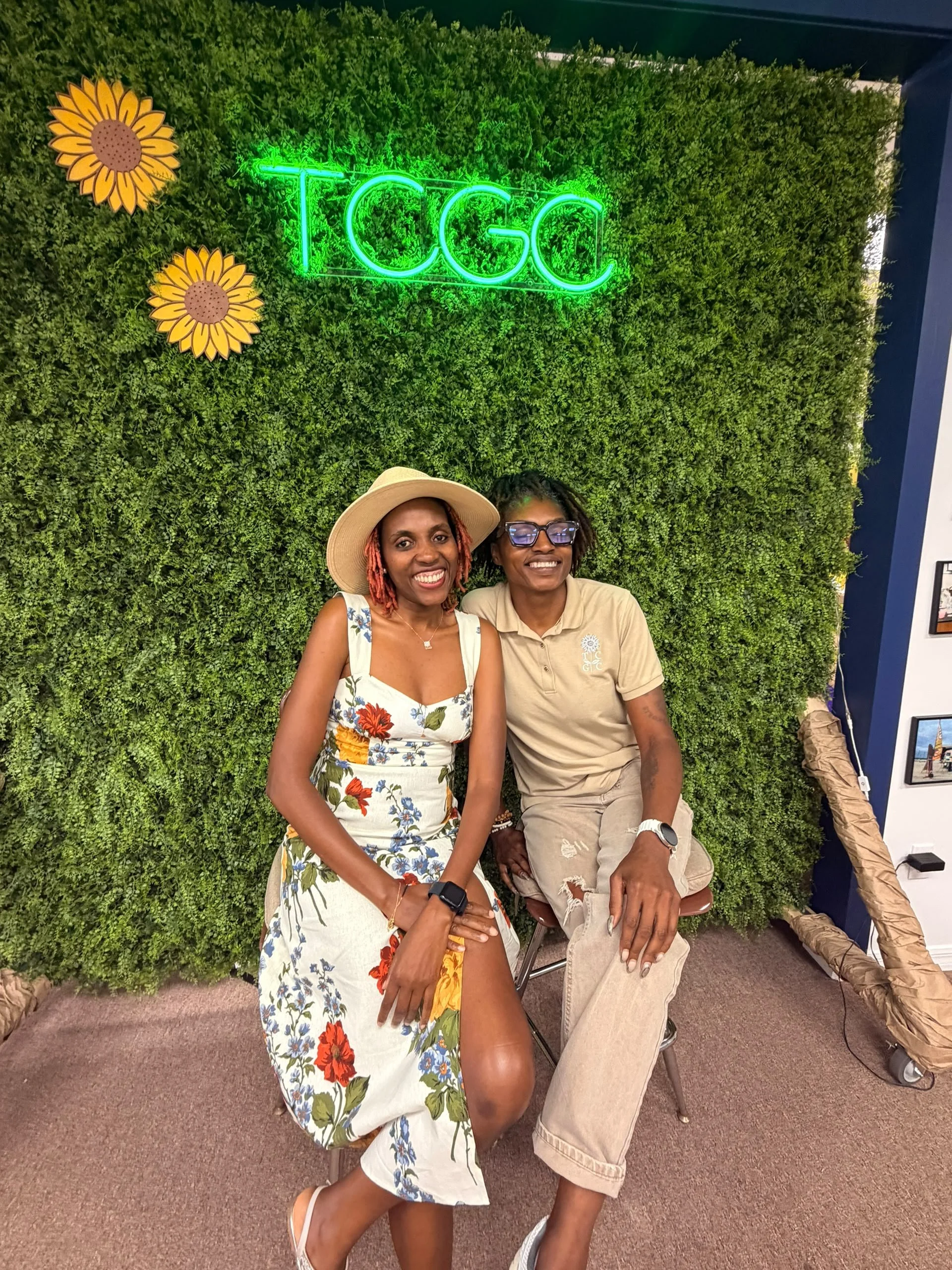 Two women sitting in front of a green wall with sunflowers and a neon sign that says