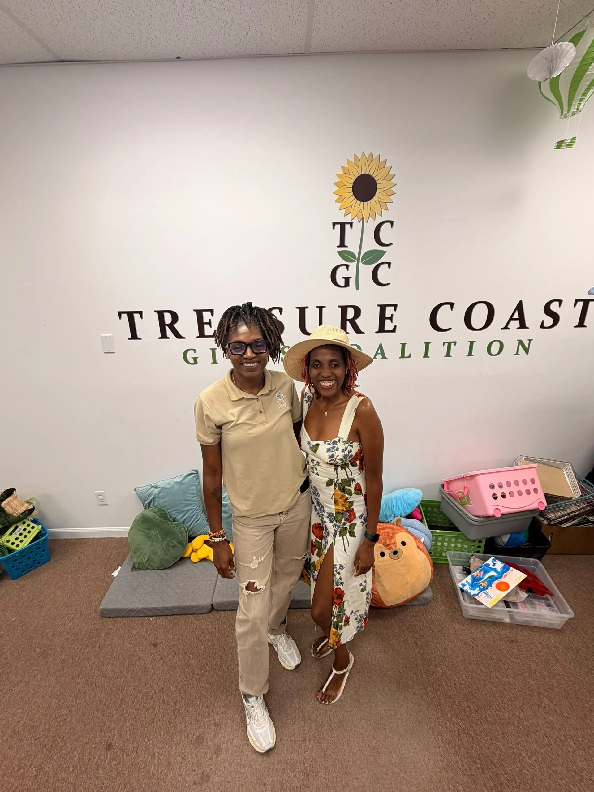Two women standing in front of a white wall with a sunflower logo and the words "Treasure Coast Growth and Education". The women are smiling, one wearing glasses and casual clothes, the other wearing a floral dress and a sun hat. There are various items and toys on the floor behind them.