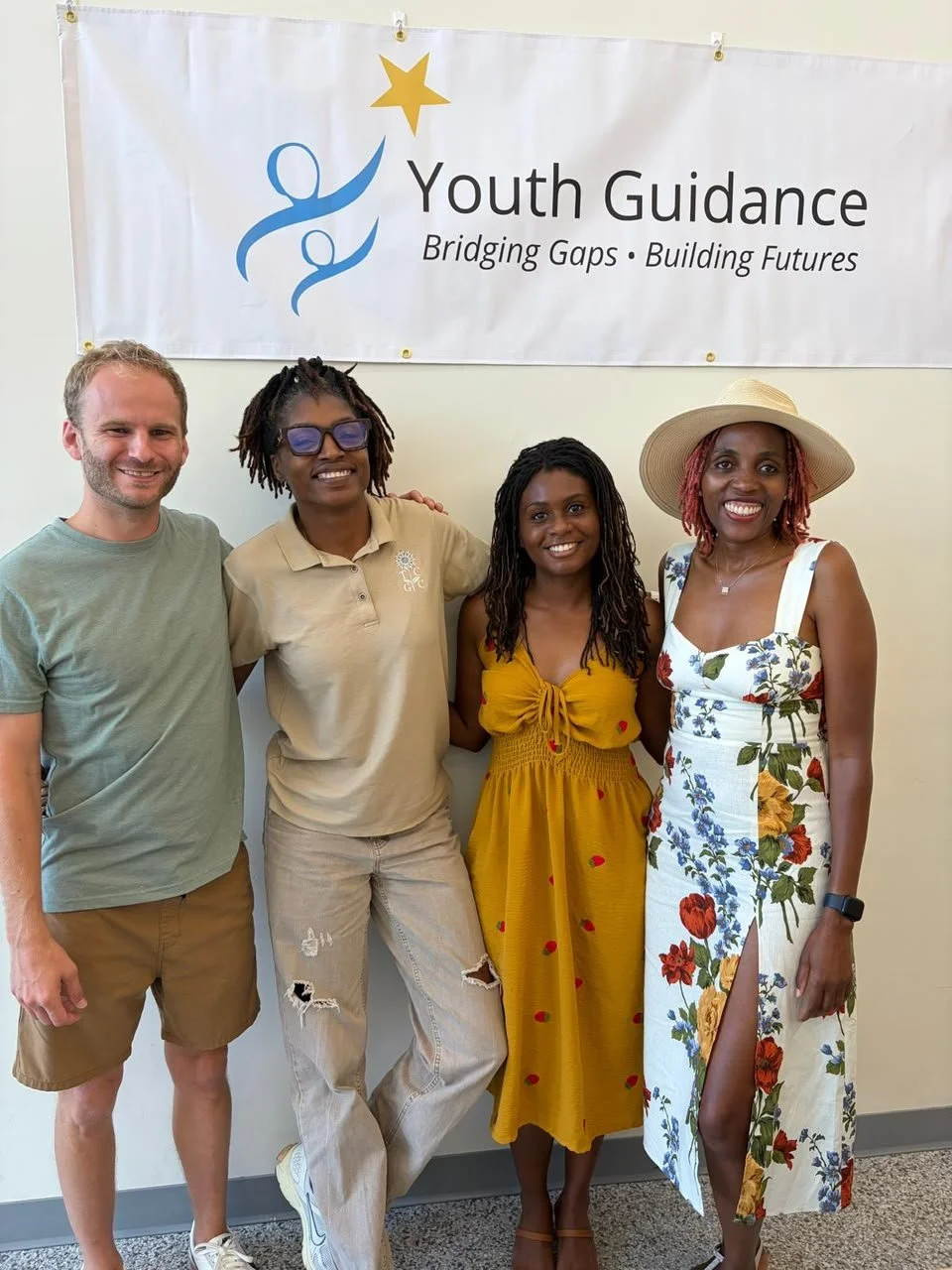 Four diverse individuals standing together in front of a Youth Guidance banner, smiling at the camera, in a well-lit indoor setting.