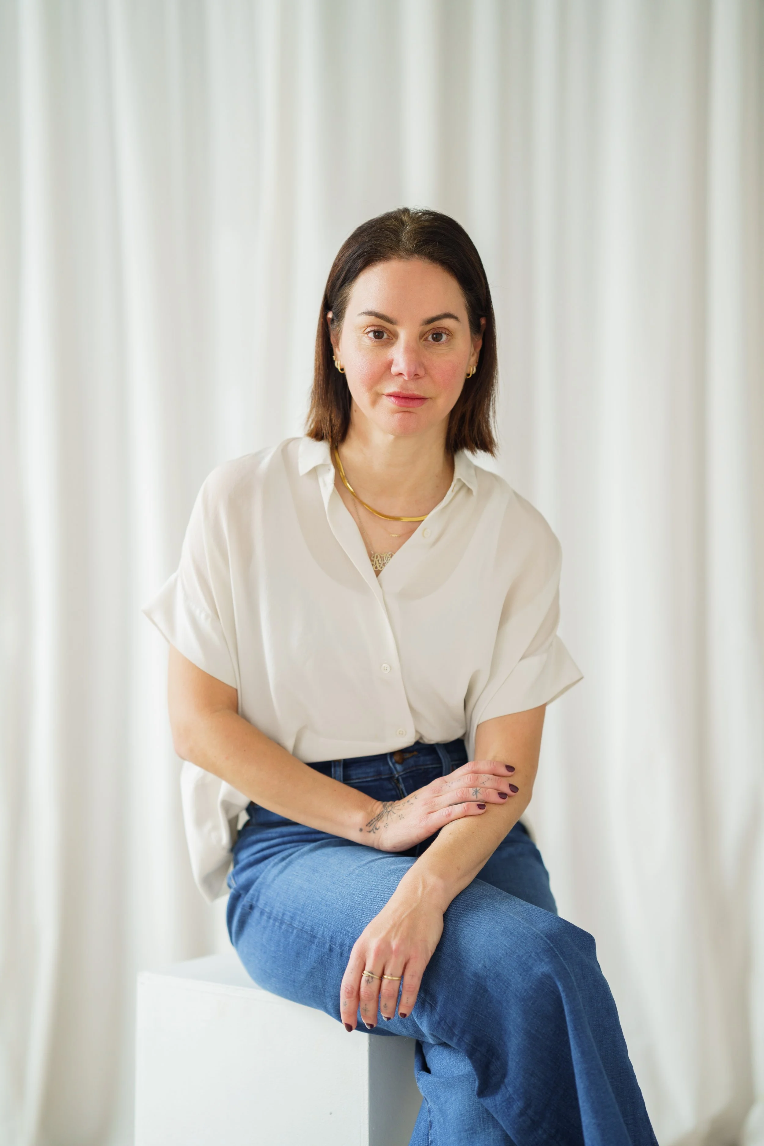 A woman with shoulder-length dark brown hair, wearing a white button-up shirt and blue jeans, sitting on a white cube in front of white curtains.