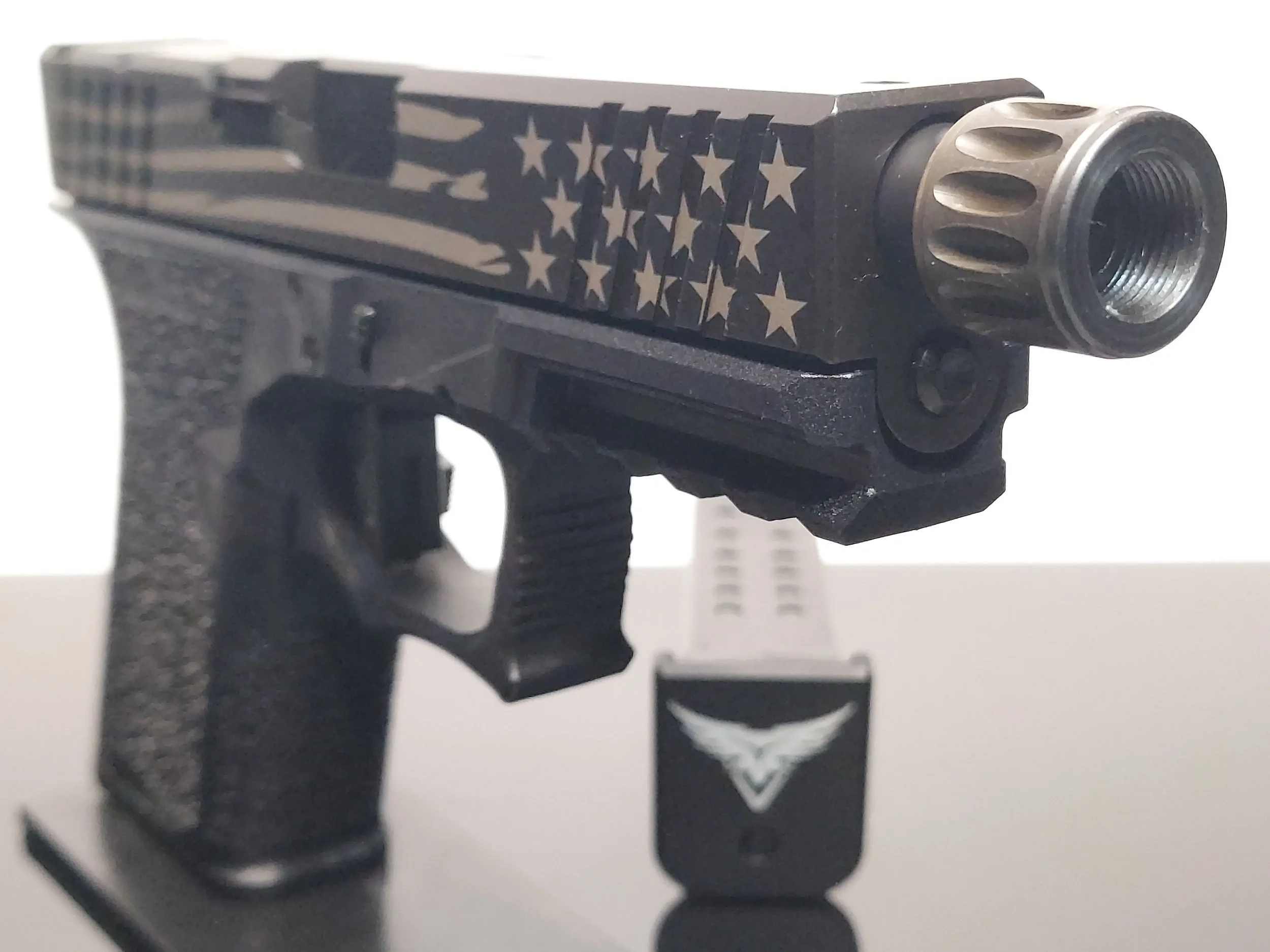 A black handgun with an American flag design and a silver barrel, with a magazine in front of it displaying a logo with a white bird.