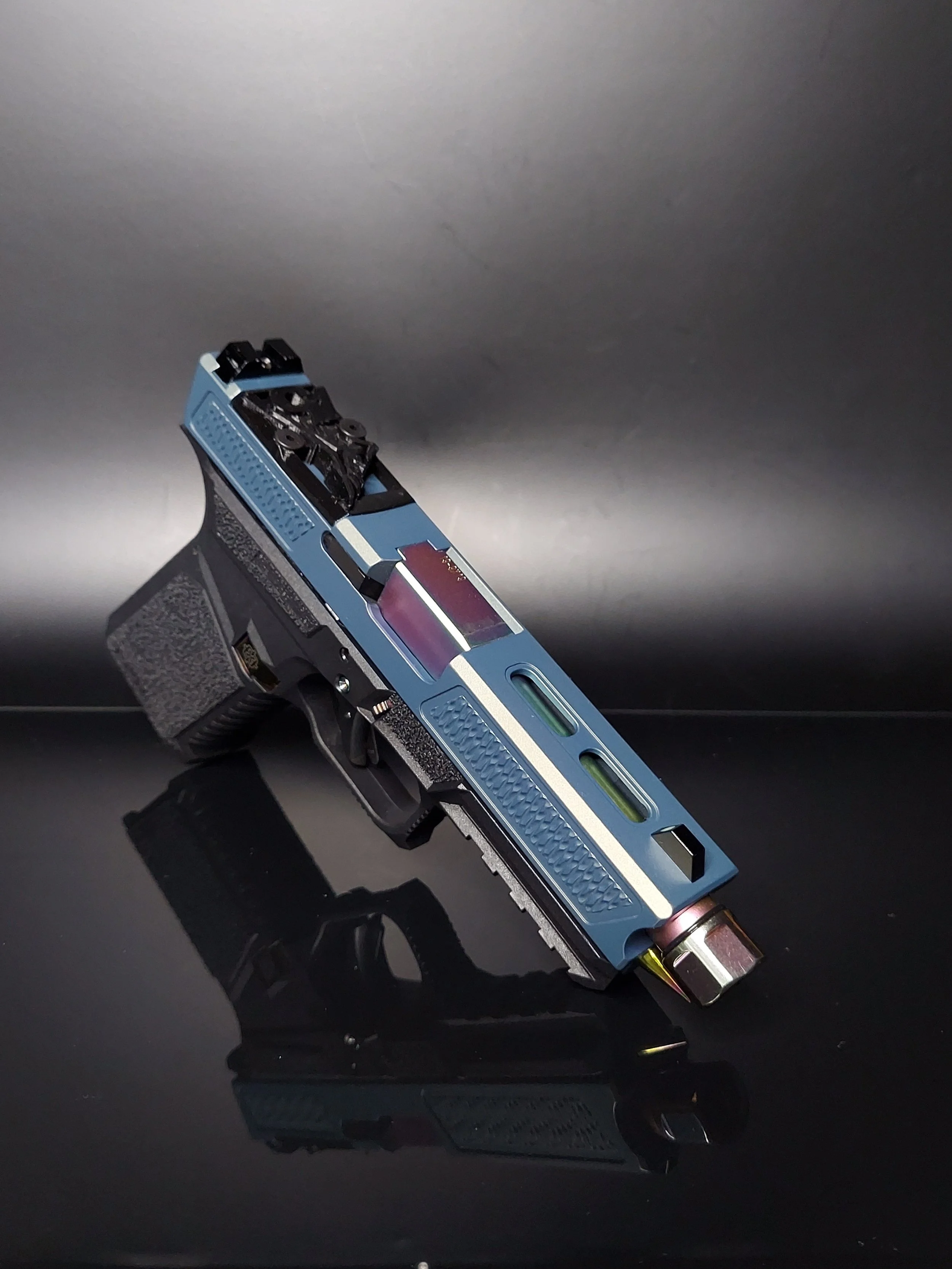 A modern, customized semi-automatic pistol with a multi-color slide, including blue, purple, and green accents, placed on a reflective black surface against a dark background.