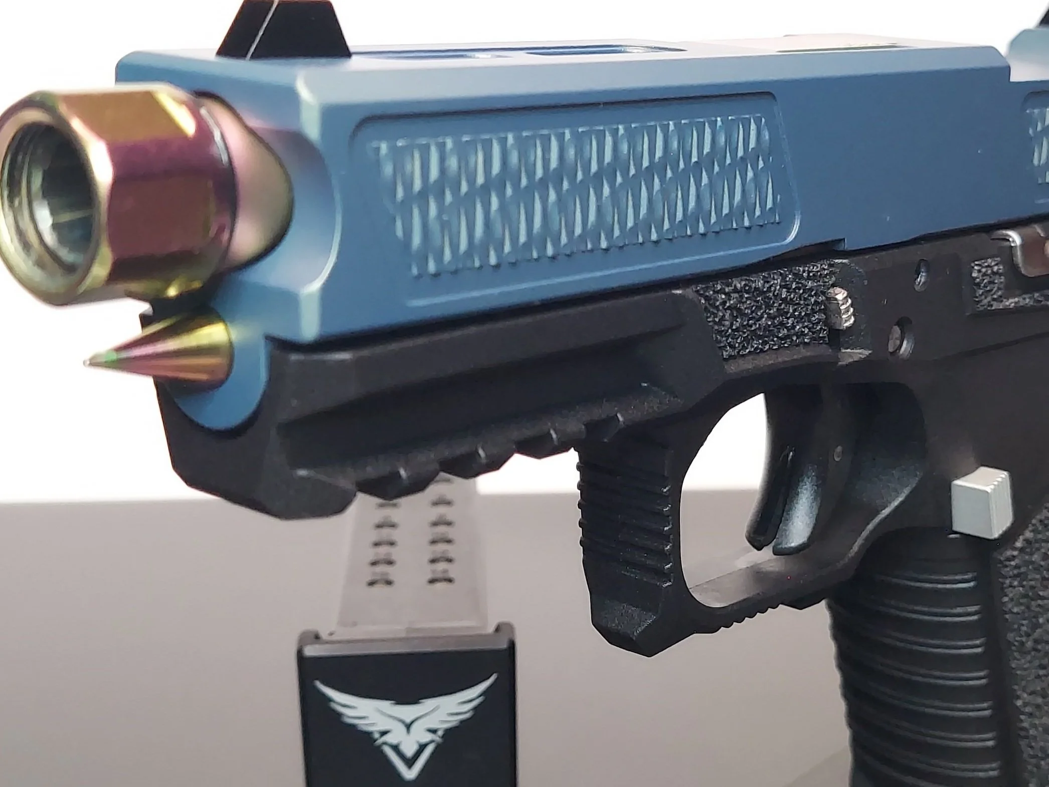 Close-up of a toy or replica gun with metallic accents and a small pin on the grip, resting against a white surface with a black and white eagle logo visible.