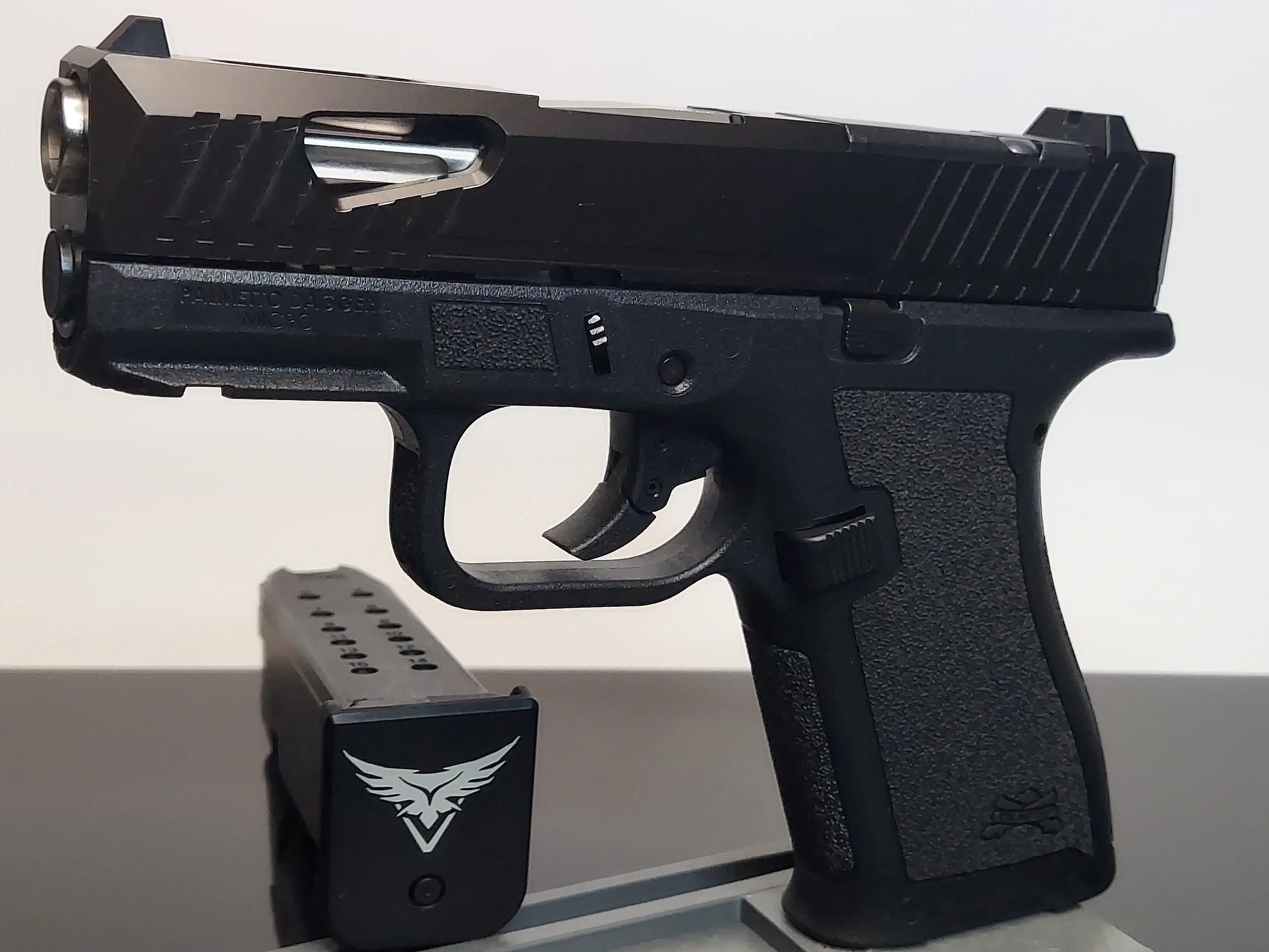 A black semi-automatic handgun with a textured grip, slide, and trigger, resting on a gray surface. The gun is fitted with a magazine featuring a white emblem of wings and a V-shape.