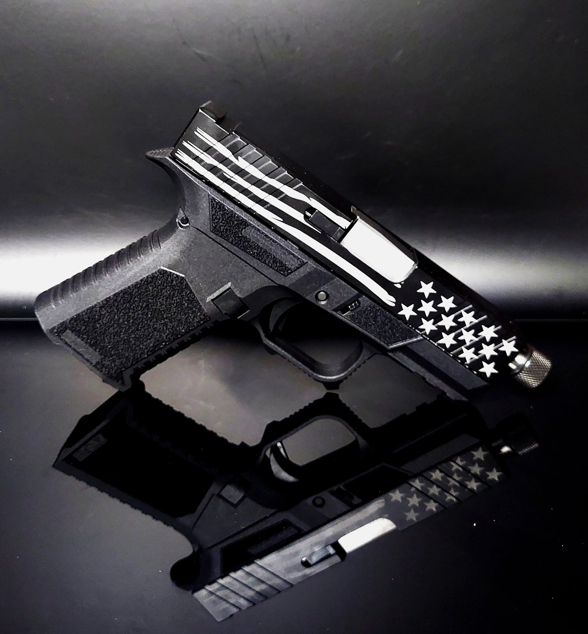 A black handgun with a star-spangled American flag design on the slide, resting on a reflective black surface.