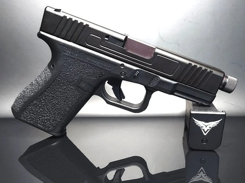 A black semi-automatic handgun resting on a reflective surface with a small black device with a white winged emblem attached near the trigger.
