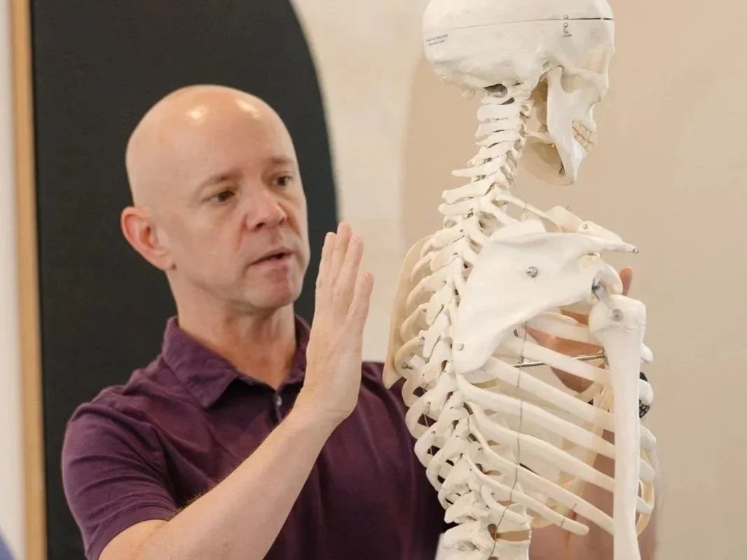 A man is explaining anatomy with a human skeleton model in a classroom or educational setting.
