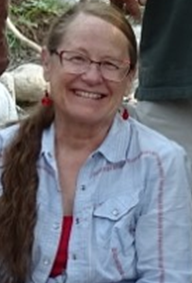 Close-up of smiling face. Long hair in a ponytail, glasses, red earrings, and a blue shirt.