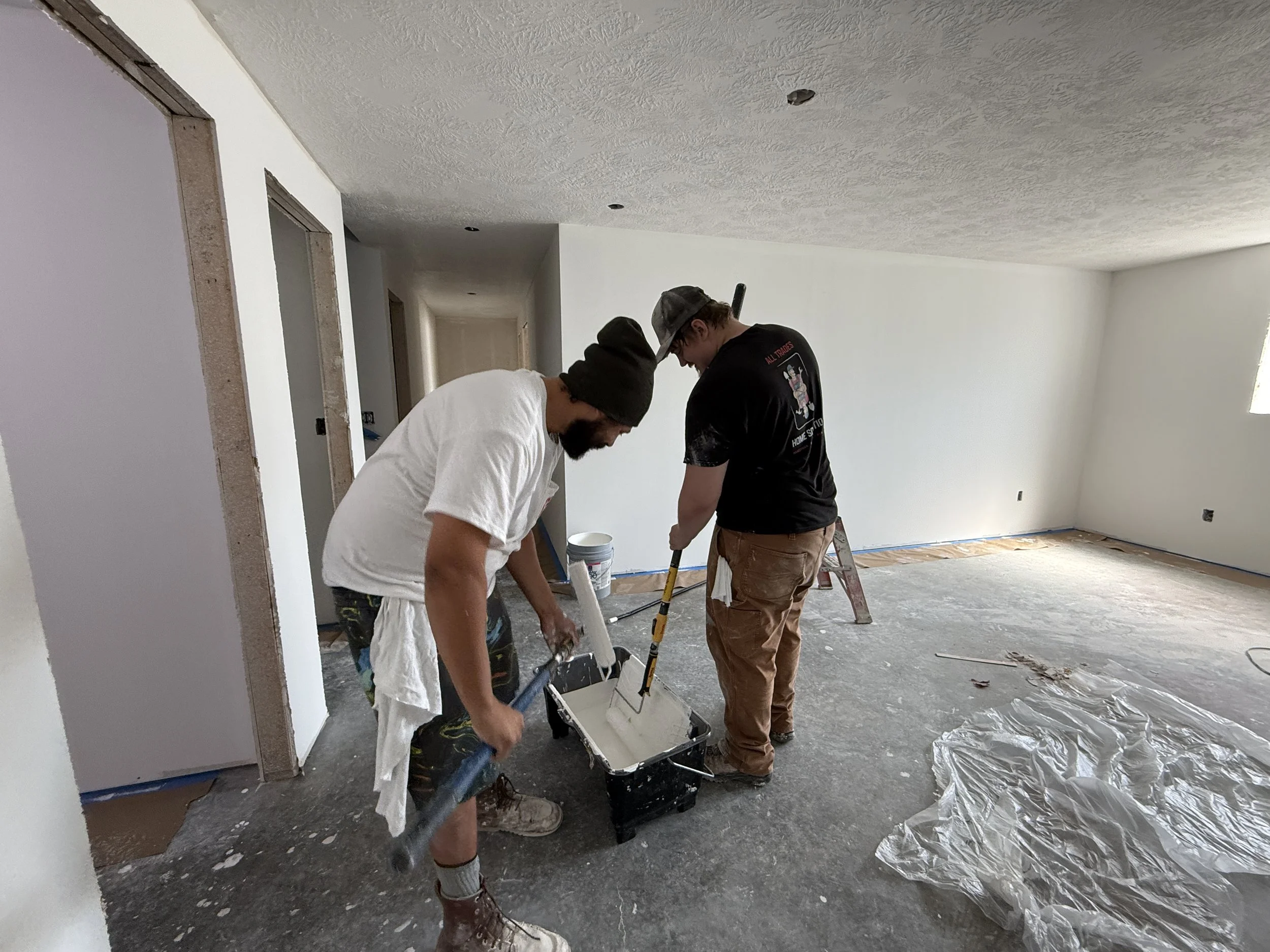 Two men are working together painting the walls of a room. One is using a roller to apply white paint, and the other is holding a paint tray. The room is under renovation, with plastic sheeting on the floor and unfinished doorways.