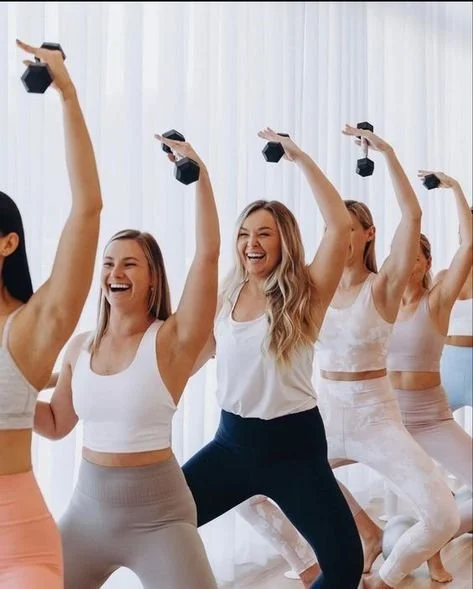Group of women exercising with dumbbells in a barre fitness class, wearing workout clothes, looking happy.