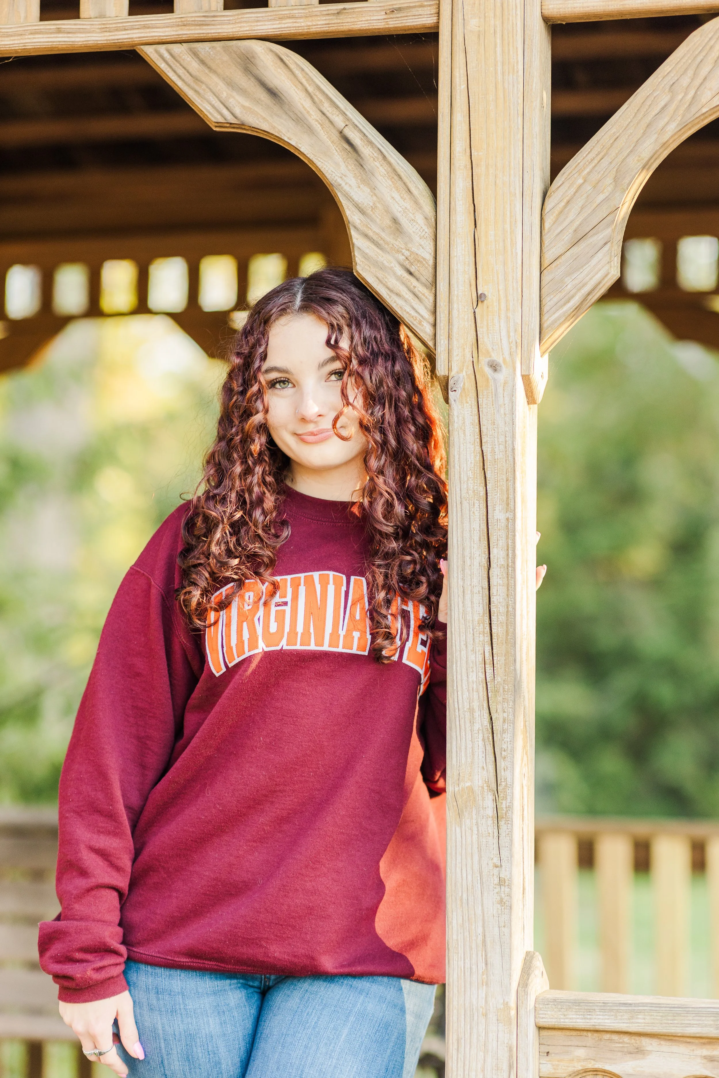 A young woman with curly reddish hair standing outside, hiding partly behind a wooden post of a gazebo or porch, wearing a maroon Virginia Tech sweatshirt and blue jeans, with greenery in the background.