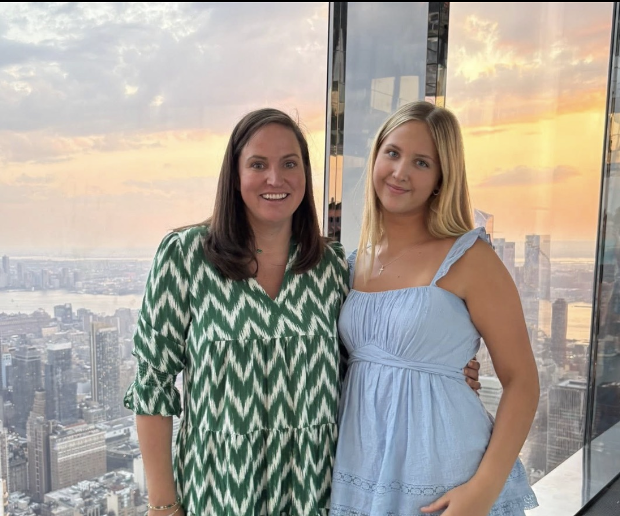 Two women standing together on a balcony at sunset with city skyline in the background. One woman is wearing a green and white patterned dress, and the other is wearing a light blue dress. They are smiling at the camera.