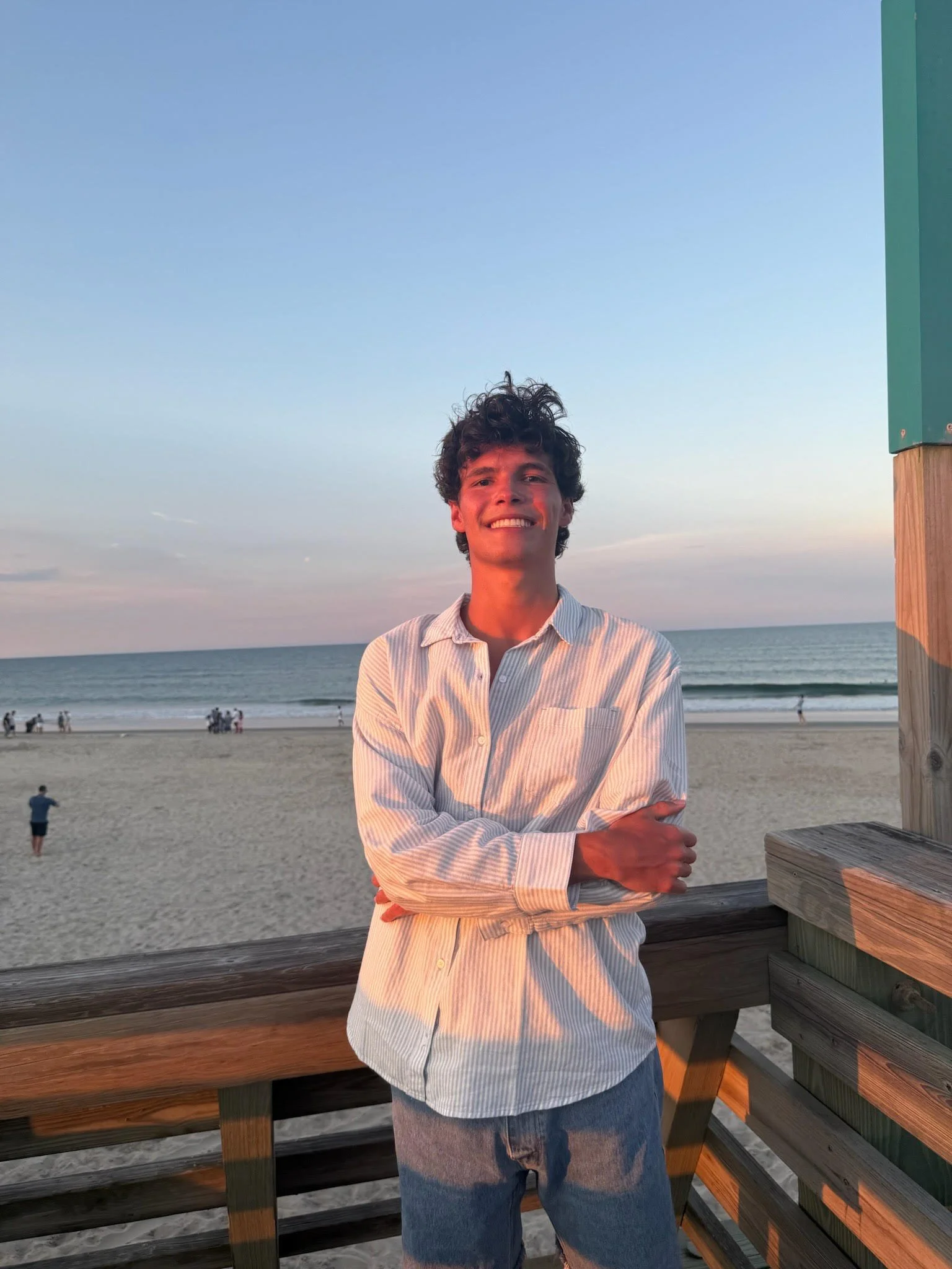 A young man with curly hair smiling, standing on a beach during sunset with the ocean in the background, wearing a striped shirt and jeans.