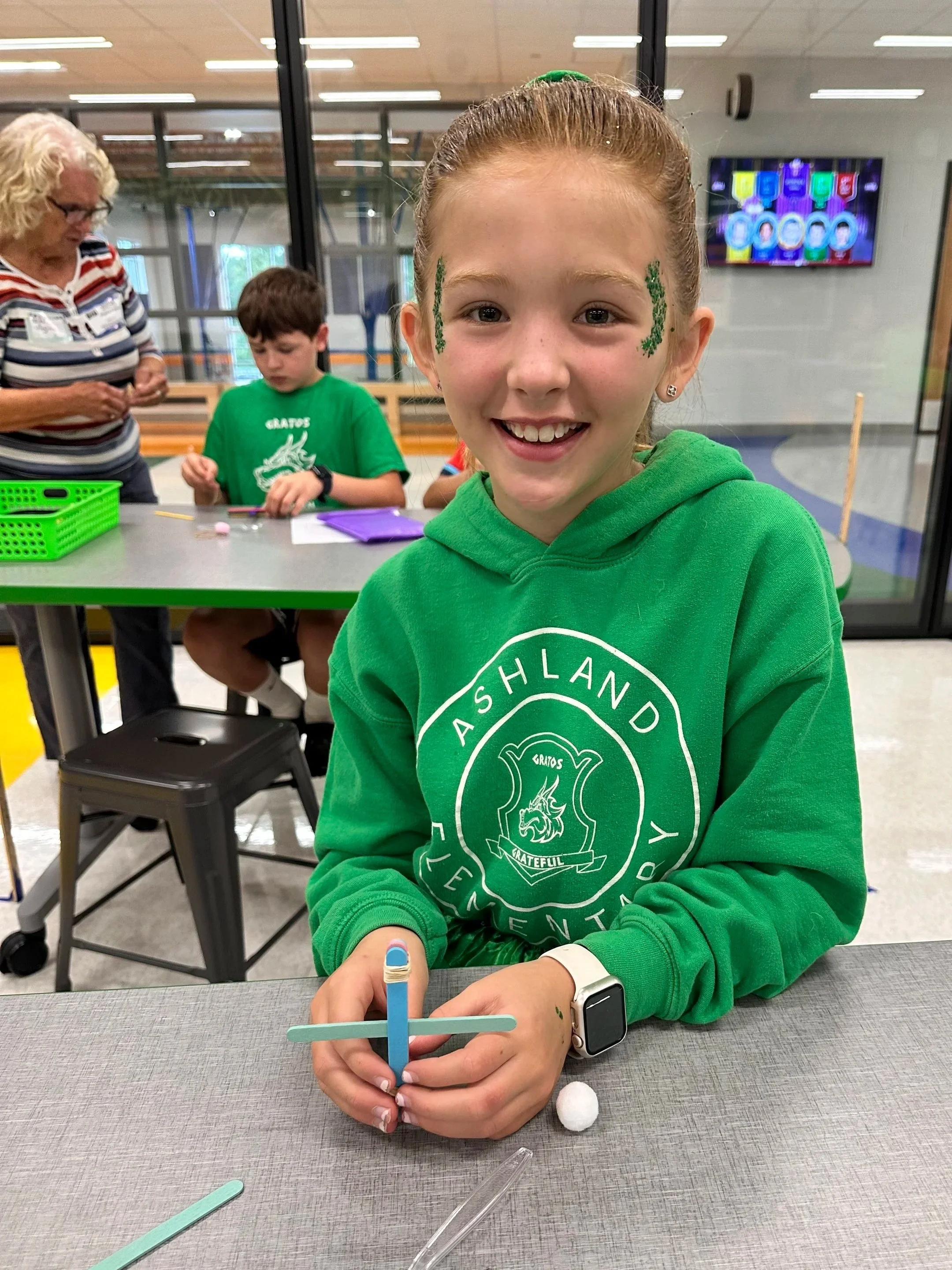 A young girl with red hair smiling at the camera, sitting at a table, holding a craft stick in her right hand. She is wearing a green hoodie with a logo on it, a smartwatch, and had face paint on her cheeks. In the background, there are two boys and an older woman engaging in a craft activity in a room with glass walls and a TV screen.