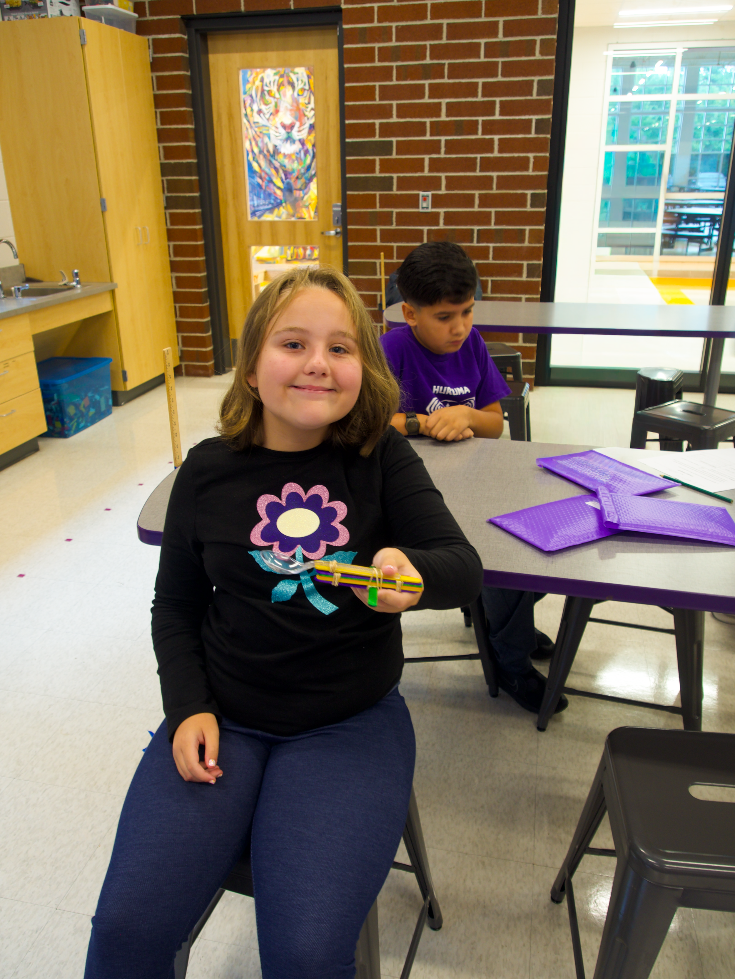 A girl sitting at a desk in a classroom, holding a large rubber band, with a boy sitting behind her. The classroom has brick walls, cabinets, and a door with a colorful tiger painting on it.