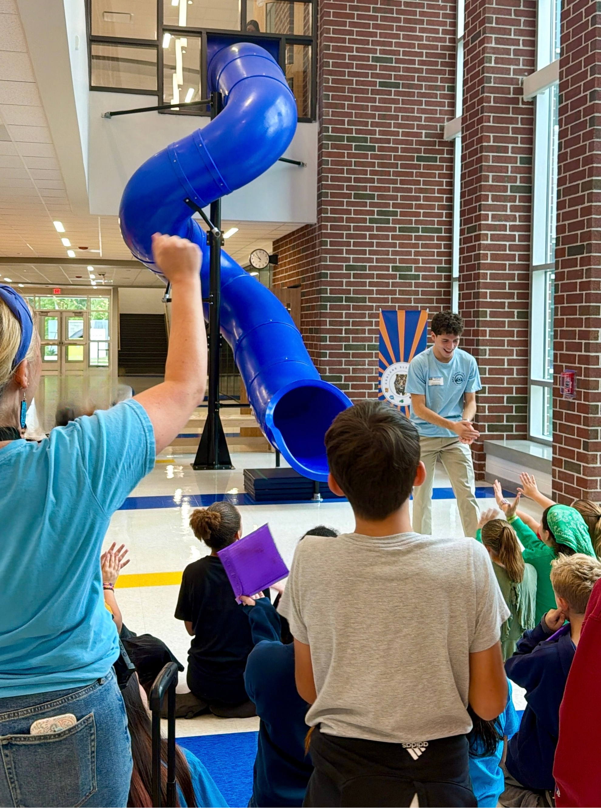 Children and adults watching a presentation in a large indoor space with a blue slide used for entertainment. A smiling young man is presenting; an audience of children is seated on the floor, raising their hands.
