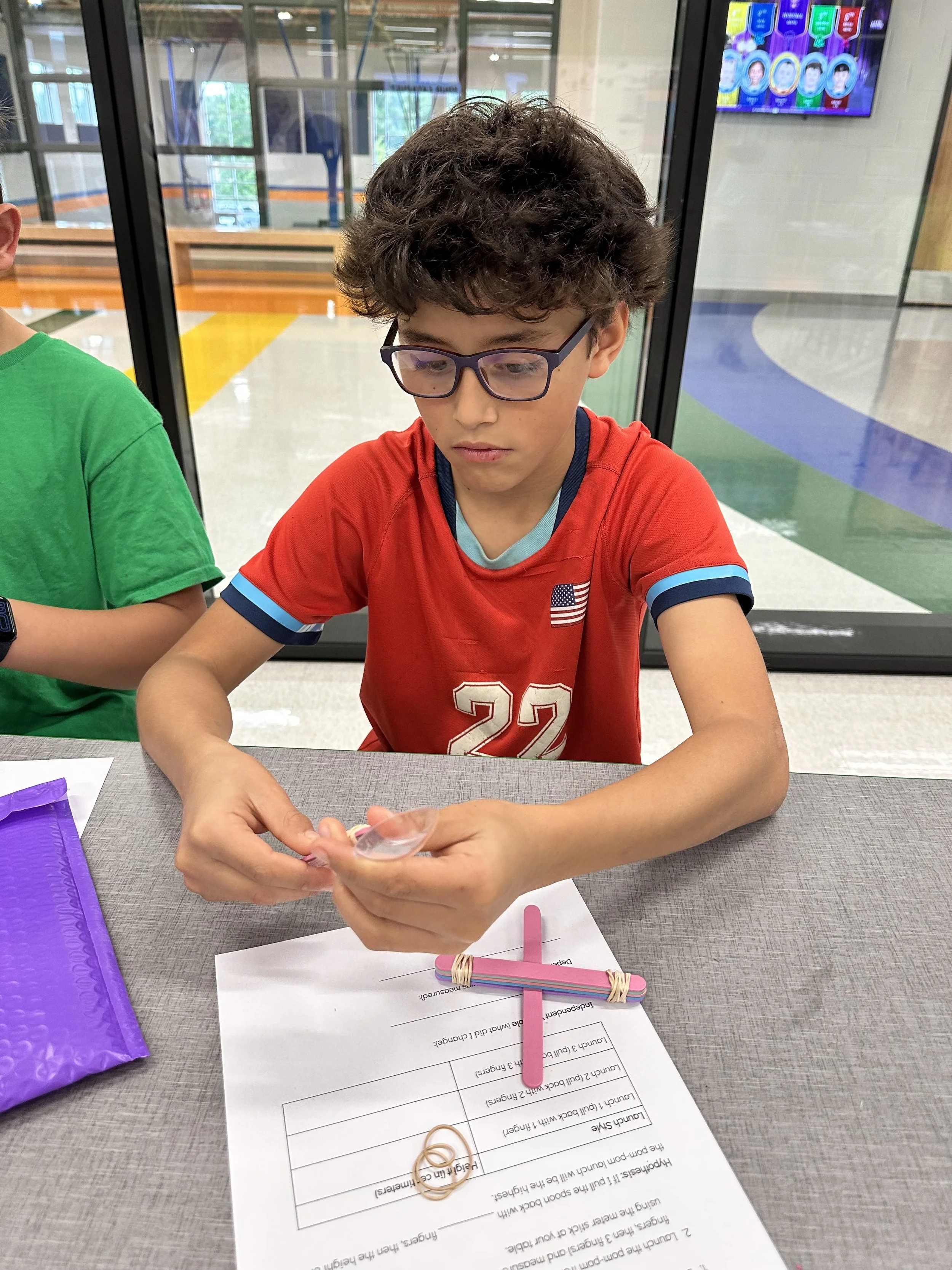 A boy with curly brown hair and glasses wearing a red t-shirt sitting at a table, holding a rubber band. A paper with questions and a pile of colorful popsicle sticks are on the table in front of him. To his left, part of another child wearing a green shirt is visible. The background shows a glass wall with a view of the indoor wakeboard area and a television screen displaying cartoon faces.