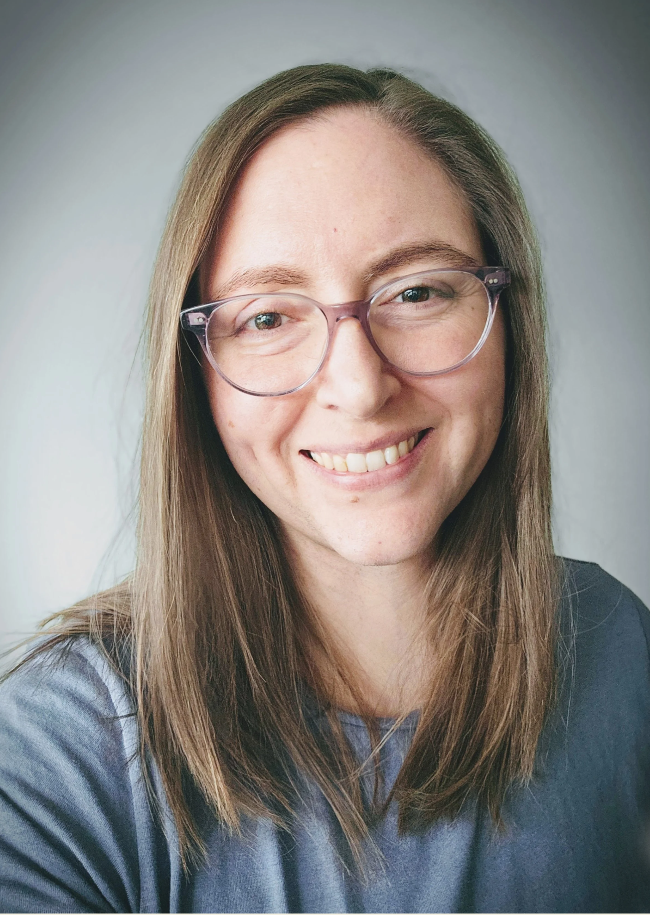 Close-up of a smiling woman with long brown hair, wearing clear glasses and a gray shirt, against a plain light background.