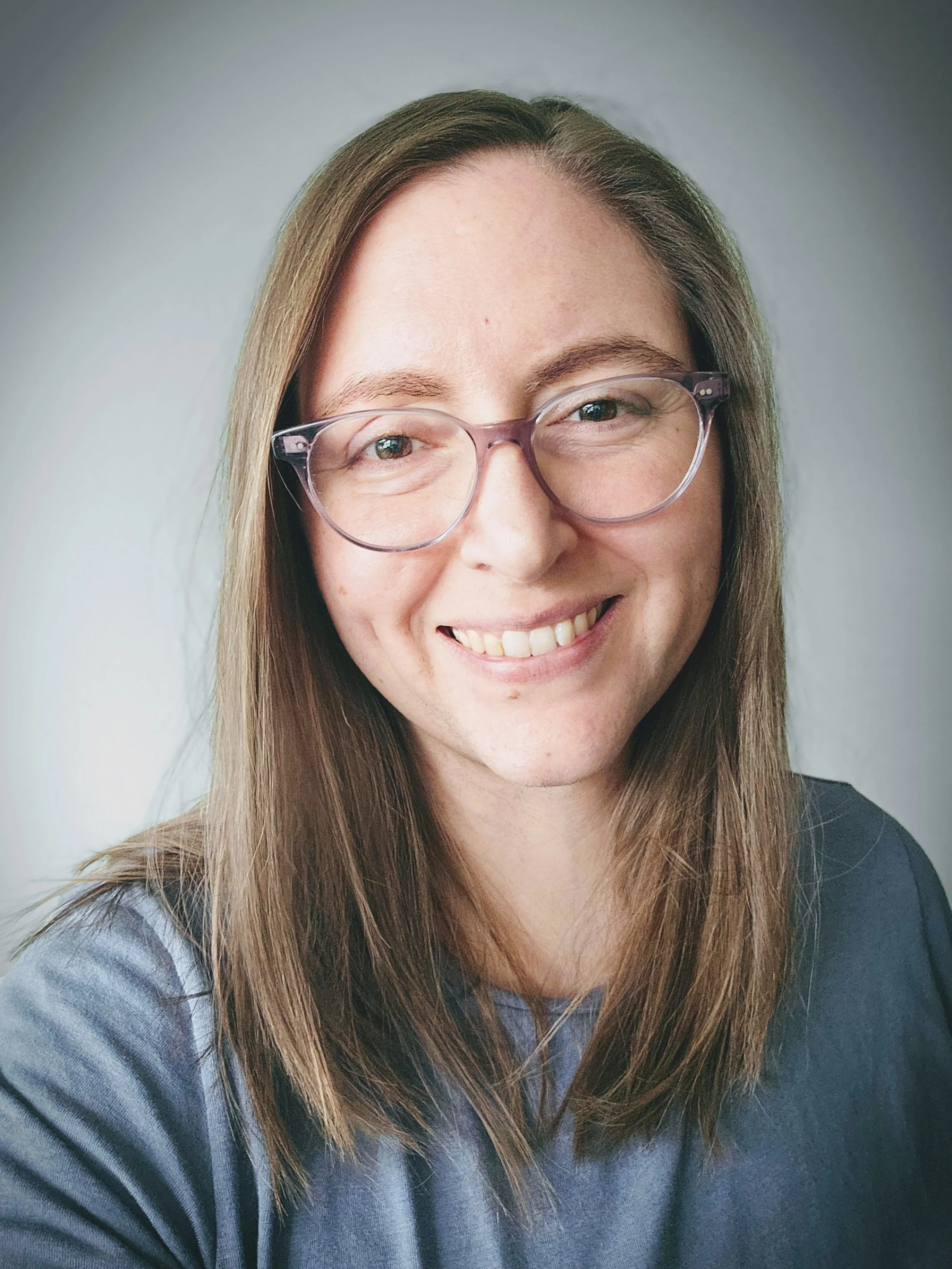 A young woman with long light brown hair, wearing glasses with pink frames, smiling, and wearing a blue shirt.