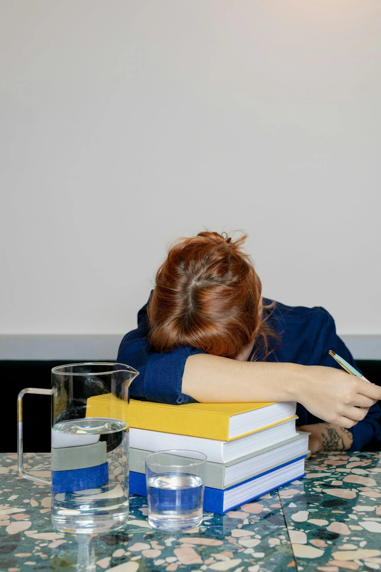 A person with red hair resting their head on a table covered with a patterned tablecloth, surrounded by books and glass pitchers of water.