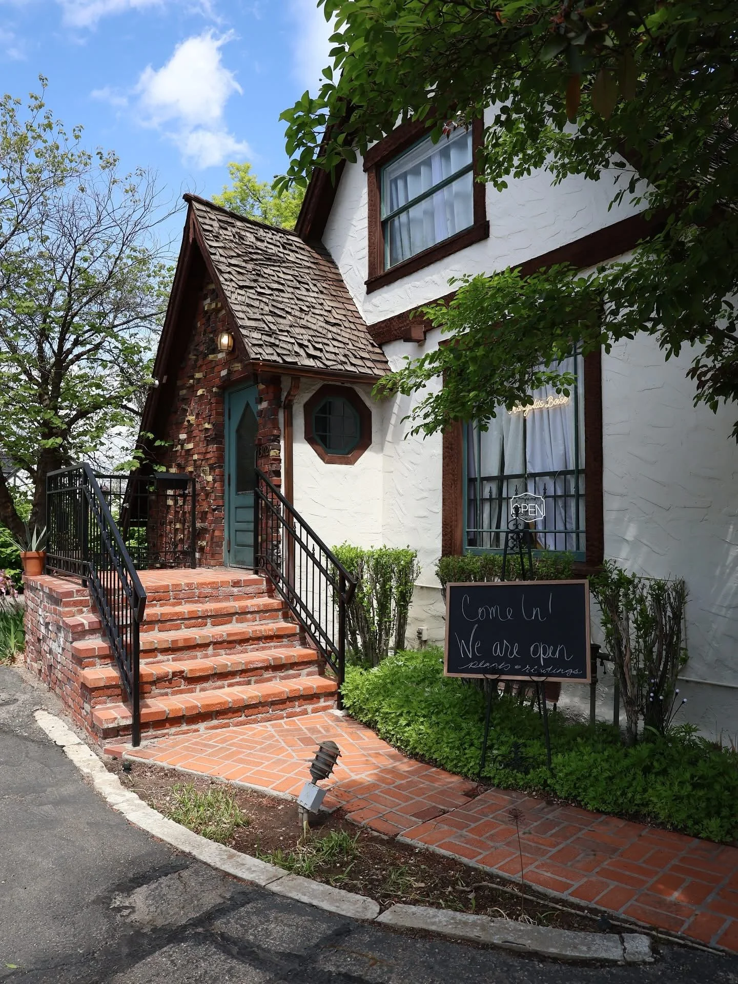 A white house with brown trim and a small brick staircase leading to a door. There is a blackboard sign outside that says 'Come in! We are open' and a window with a neon 'Open' sign. Trees and bushes surround the building under a partly cloudy sky.