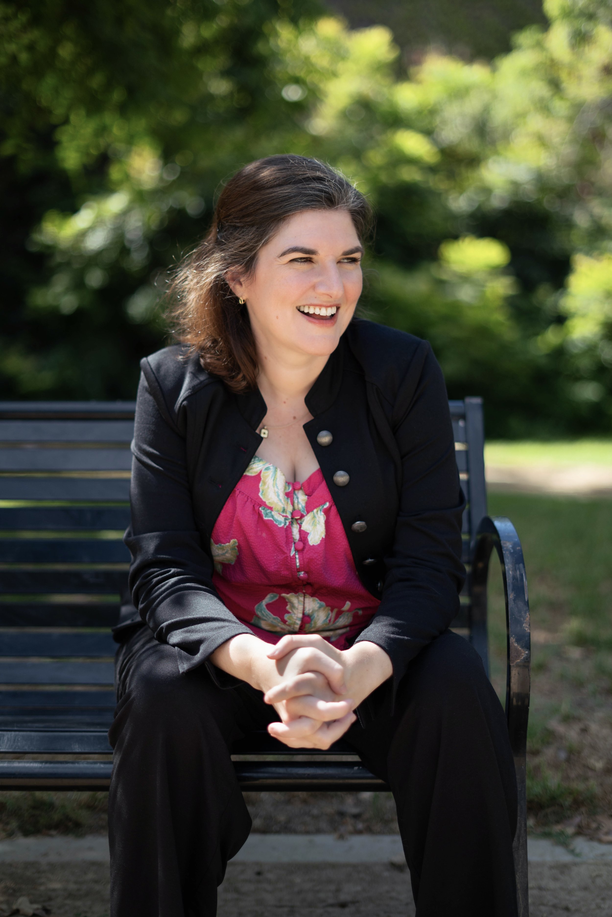 A woman sitting on a park bench, smiling, with blurred green trees in the background.