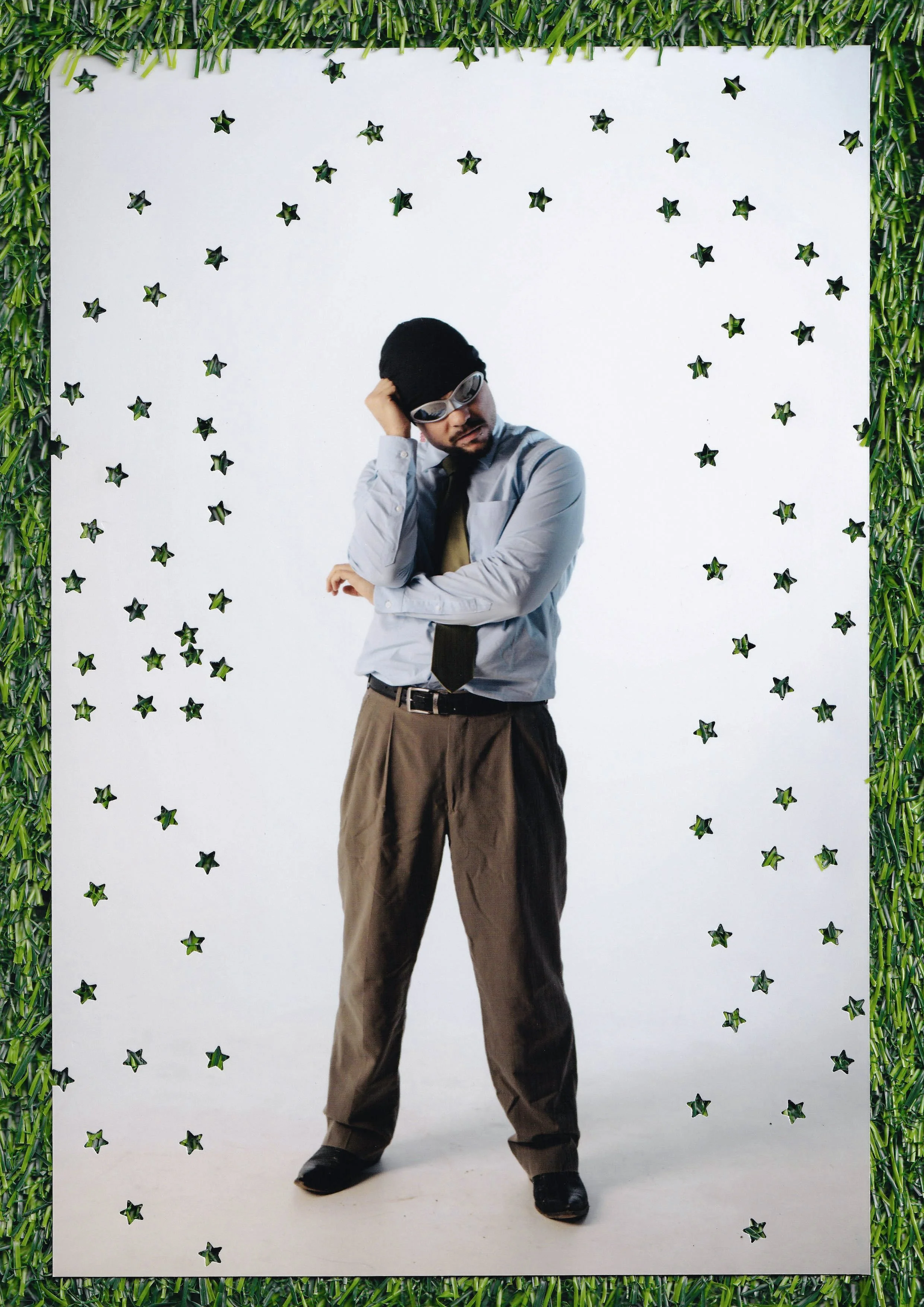 A man wearing sunglasses, a light blue shirt, a black tie, and brown pants stands against a white background decorated with small black star-shaped cutouts. He is holding his head with one hand and appears to be posing thoughtfully.