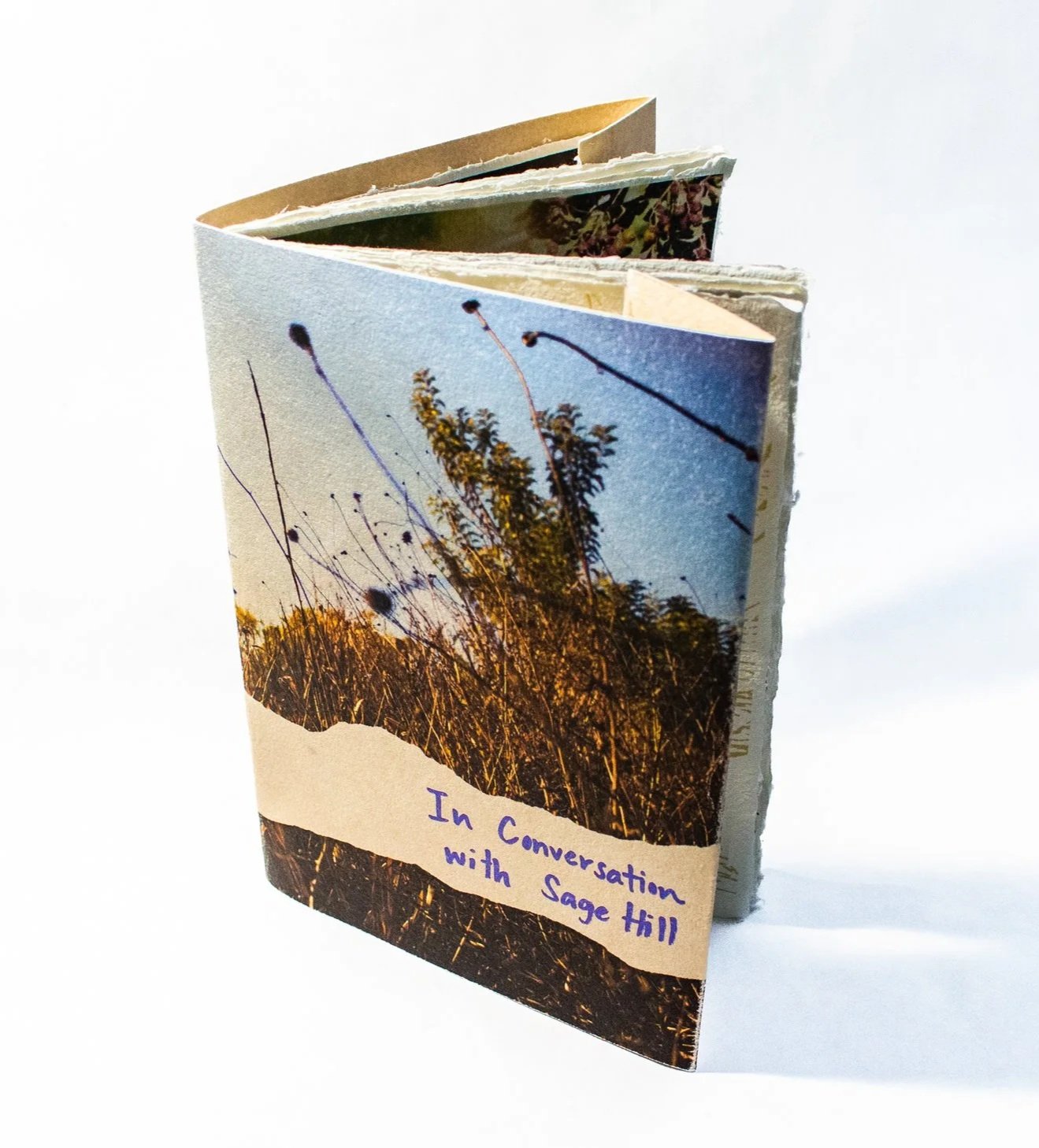 A small open book with a covers showing a field scene with tall grass and trees under a blue sky. The title handwritten on paper label on the cover reads "In Conversation with Sage Hill."